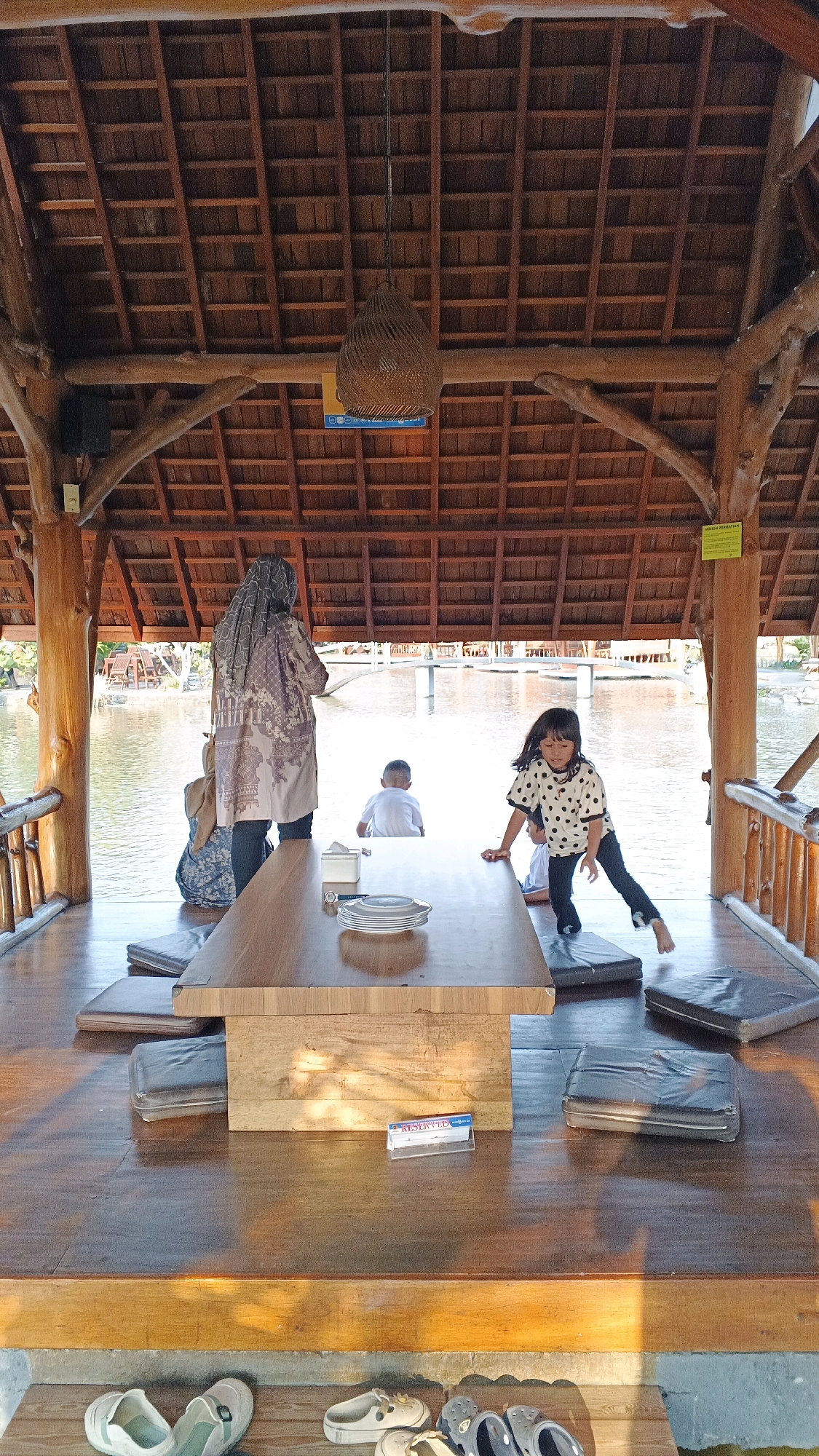 A casual outdoor dining setting in Minggir, Indonesia, is depicted with a view of a body of water. The foreground features a wooden table with a stack of plates and a "RESERVED" sign. To the left, a woman wearing a hijab and a patterned tunic stands near a blue backpack. In the mid-ground, a young boy in a white shirt sits facing away from the camera, and to his right, a girl with dark hair, wearing a black and white polka-dot shirt and black pants, is in mid-motion, leaning over the table. She appears to be either stepping onto or off a dark mat. Several dark cushions are arranged on the wooden floor around the table. The background reveals a calm body of water with some structures and vegetation visible in the distance. Above, a woven light fixture hangs from the intricately constructed wooden ceiling, supported by thick, natural wood beams. The overall ambiance suggests a relaxed dining experience in a tropical environment.