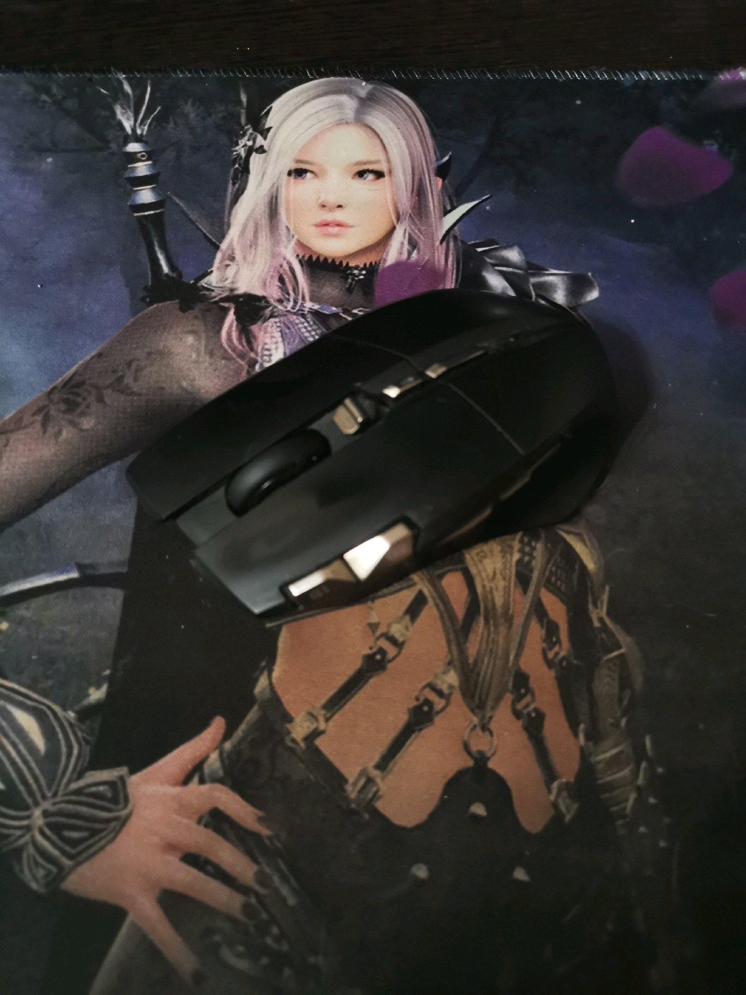 A black computer mouse is placed on a dark mousepad featuring a digital illustration of a woman with pale skin, long white hair with pink streaks, and dark eye makeup. The woman is depicted wearing dark, ornate armor and jewelry. A human hand with manicured dark nails is visible on the left side of the image, resting on the mousepad. The background of the mousepad is a dark, abstract scene with purple circular shapes. The scene appears to be indoors, likely on a flat surface such as a desk. There is no indication of the city of Kemerovo, Russia, or any specific urban location within the image.