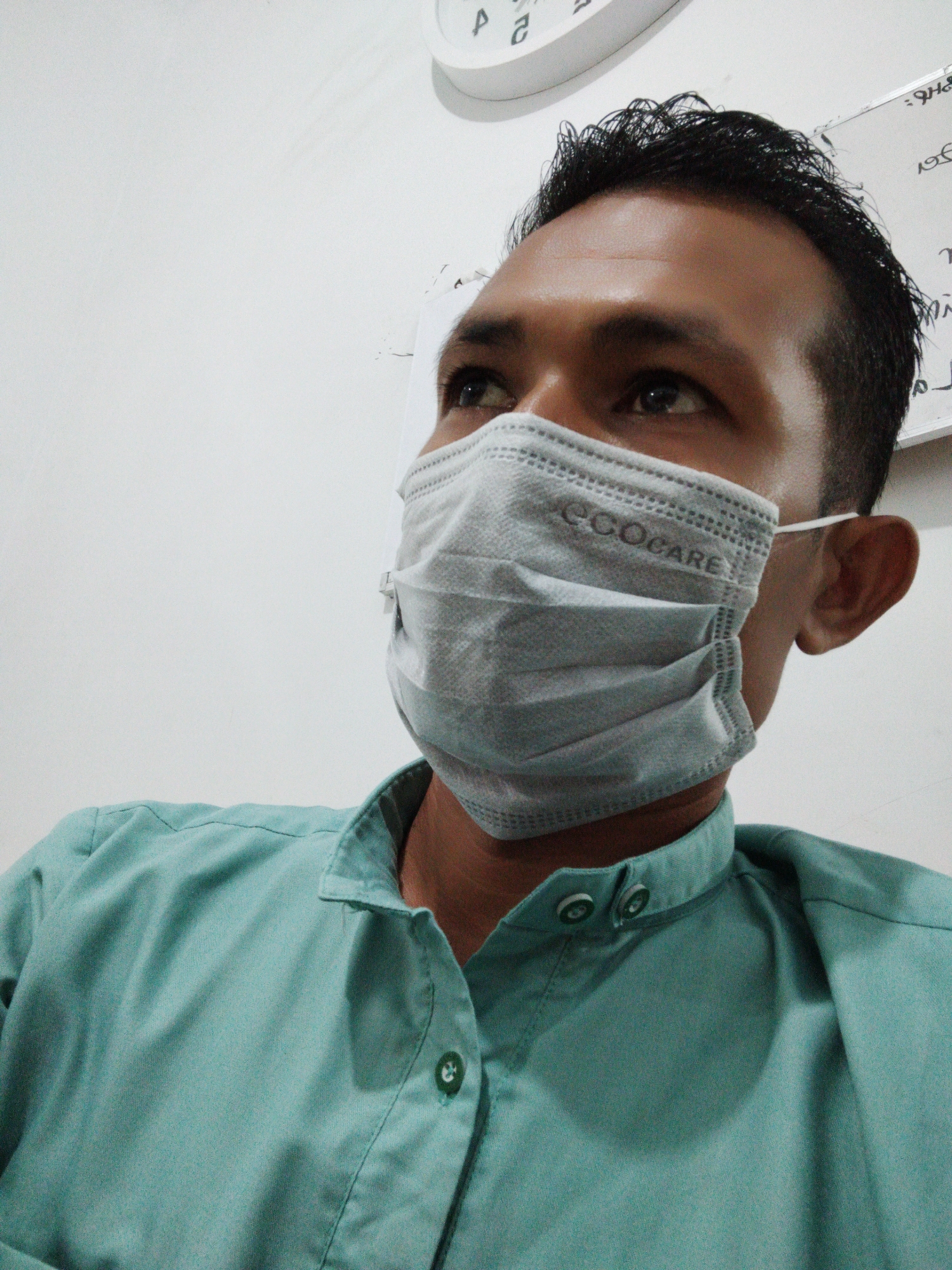 A person is wearing a light blue shirt and a gray face mask with the word "ECOCARE" printed on it. They are looking up and to the left. The background is a white wall with a clock and some writing on it. The photograph was taken indoors, location in the city of Lhokseumawe, Indonesia is unknown.