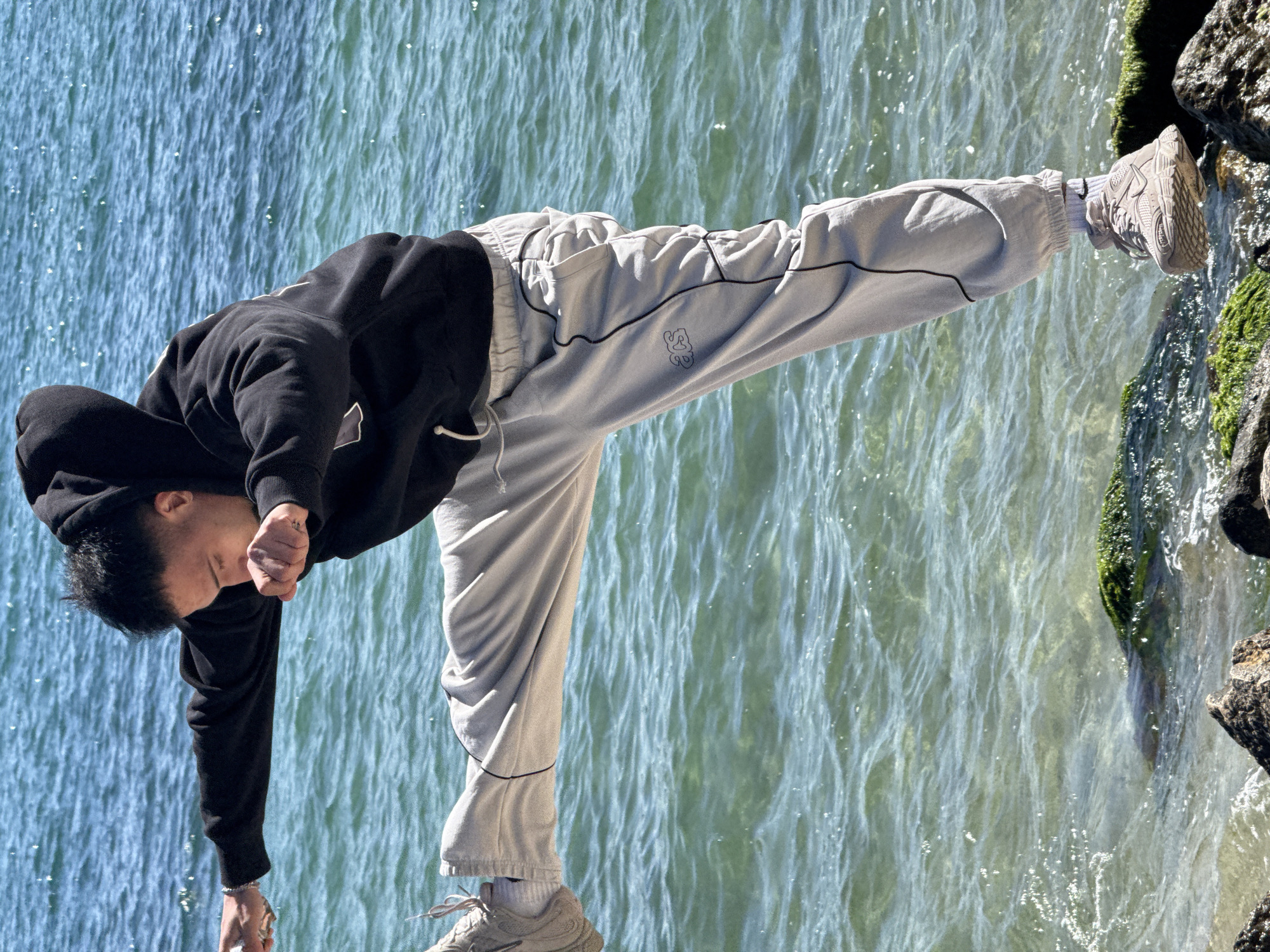 The image shows a young male in a dynamic, balancing pose by a body of water in Varna, Bulgaria, under bright, sunny conditions. The subject is wearing a black hooded sweatshirt, light grey sweatpants with black piping, white socks, and light grey athletic shoes. He is balancing on his left leg, with his right leg extended horizontally to the side. His upper body is bent forward, and his head is angled downwards. His right hand is near his face, while a silver ring is visible on his left pinky finger. On the left thigh of his sweatpants, there is a small, stylized logo that resembles "SG" or "SC".
The setting features a clear body of water, likely the Black Sea or a lake, which fills the majority of the background. The water's surface is rippled and shows glints of sunlight. On the right side of the frame, there are moss-covered rocks forming a natural shoreline where the person is standing. The overall atmosphere suggests an active and pleasant outdoor scene.