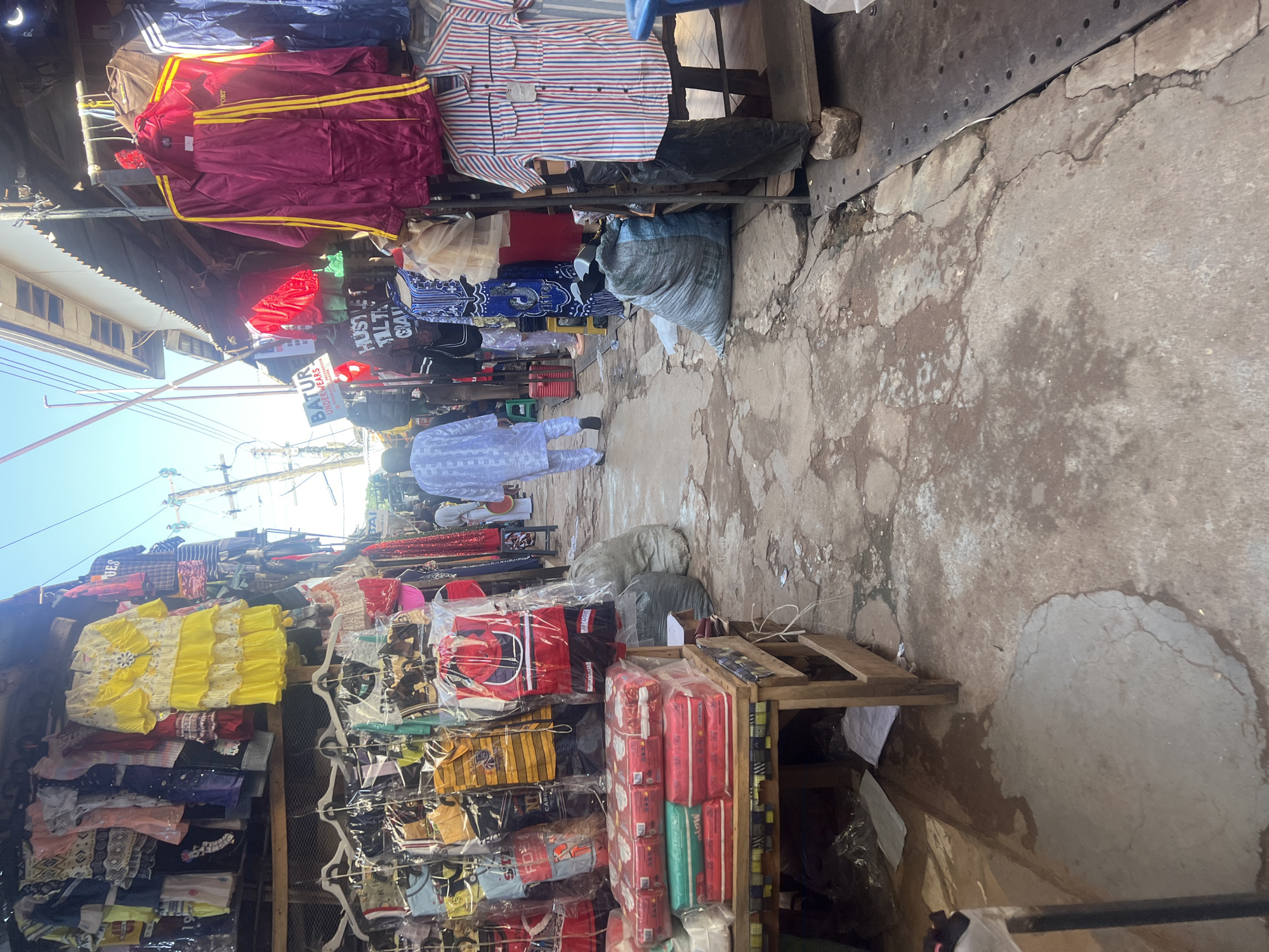 The image captures a street market scene in Jos, Nigeria, with stalls lining a narrow pathway. The stalls display a variety of clothing items, including children's wear, dresses, and tracksuits. Some garments are neatly hung, while others are packaged. In the foreground, stacks of what appear to be diapers are arranged on a wooden table. A person dressed in a white traditional outfit walks away from the camera down the market lane. Large sacks of goods are visible near the stalls, and the ground is a mix of cracked concrete and dirt. The environment suggests a bustling commercial area with limited infrastructure.