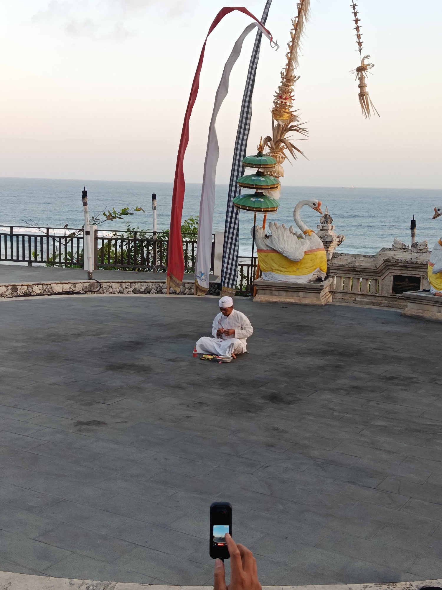 A man dressed in white sits cross-legged on a paved circular area by the ocean in Kuta Selatan, Indonesia, engaged in a ritualistic activity. He is holding a small tray with items and appears to be performing a ceremony. Behind him, a white swan statue adorned with yellow fabric stands prominently. The scene is decorated with colorful, traditional Indonesian flags and festive banners, suggesting a cultural event or religious observance. The ocean stretches out beyond a low stone wall and a railing, with a faint horizon line under a pale sky. In the foreground, a hand holds a smartphone, capturing the scene.