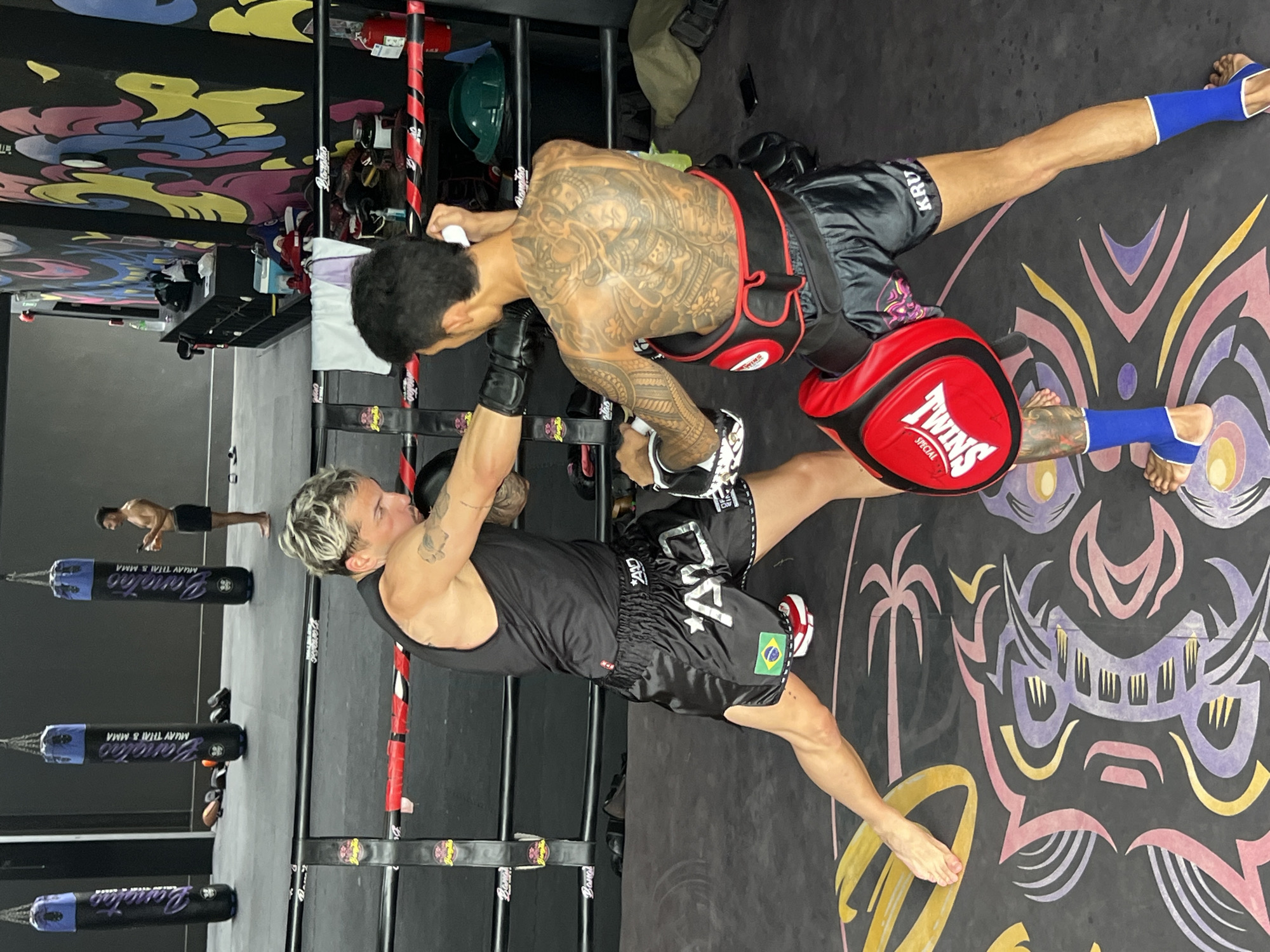 Two men are engaged in a Muay Thai training session in what appears to be a gym. The man on the left, with blonde hair, wears a black tank top and black Muay Thai shorts with a Brazilian flag patch. He is holding a red punch mitt, likely for his training partner to strike. The man on the right, with dark hair and extensive tattoos covering his torso and arms, wears black Muay Thai shorts and a red Muay Thai body protector. He is in the process of striking the mitt. Both men are barefoot.
The background reveals a Muay Thai gym setting with several tall, cylindrical punching bags hanging from a metal rack. The bags are black with purple and white text, some of which is visible as "Bangtao MUAY THAI & MMA." The floor is a dark mat with a colorful, abstract design featuring stylized figures, palm trees, and what looks like a tribal mask or demon face in purple, pink, and yellow. The overall atmosphere suggests an active training environment. The lighting appears to be indoor gym lighting, making it difficult to determine the time of day or weather. Some gym equipment, including what looks like a fire extinguisher and green helmets, are visible on shelves in the background. The text "TWINS SPECIAL" is visible on the punch mitt, indicating the brand of equipment. The overall scene depicts an intense training session focused on striking techniques.