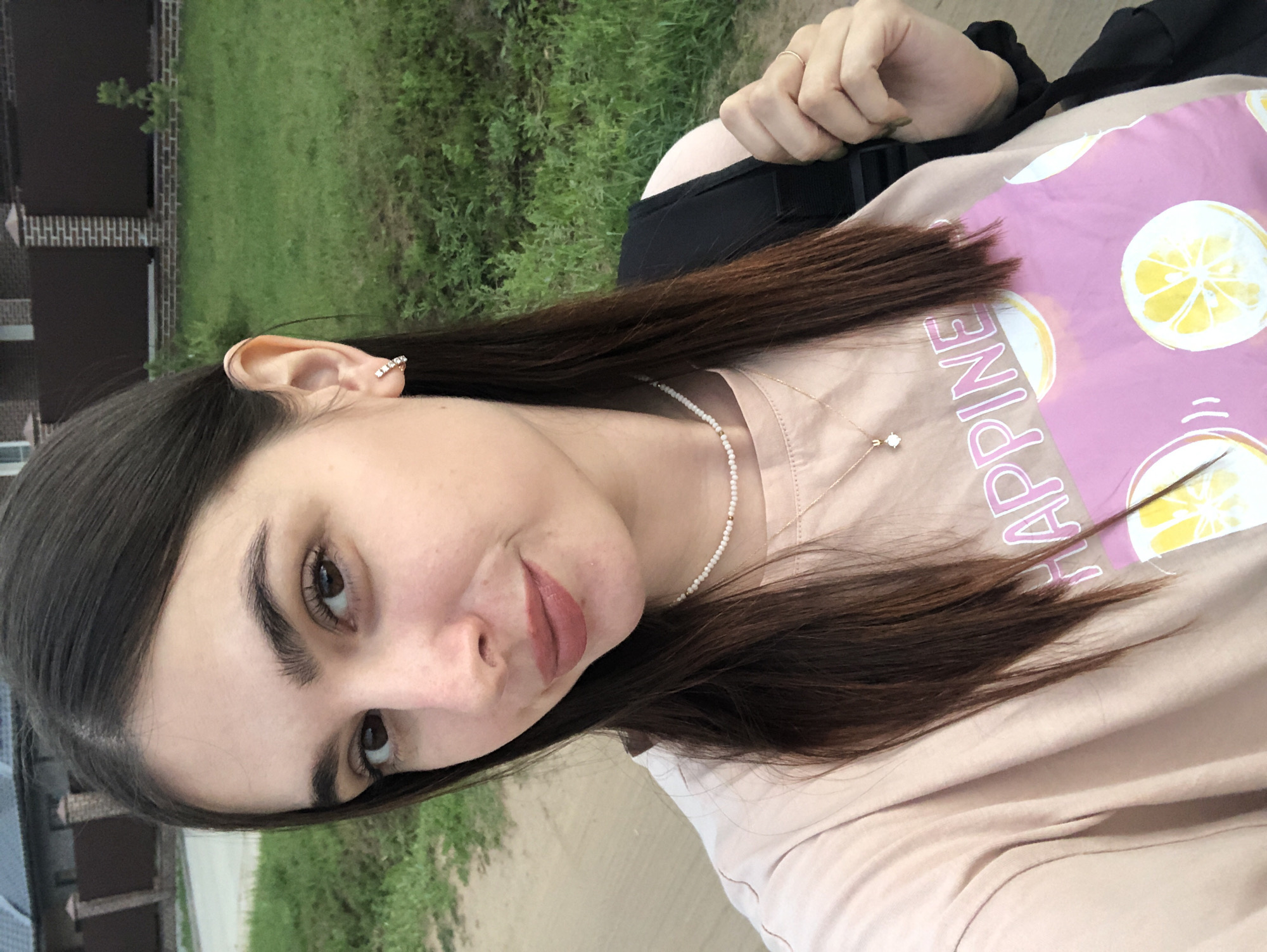 A young woman with long, dark brown hair and fair skin is pictured in a close-up selfie. She is wearing a light pink t-shirt with a graphic print of lemons and the word "HAPPINESS" in pink lettering. She has on a delicate pearl necklace and a gold pendant necklace. Her right ear is adorned with a small, sparkling earring. She has a light application of lipstick. She is holding onto the strap of a black backpack with her right hand, on which she wears a thin gold ring. The background appears to be an outdoor setting, possibly a suburban area. A paved pathway and a grassy area with a brick fence and part of a building are visible. The overall scene suggests a casual outdoor activity in Noviy, Russia.