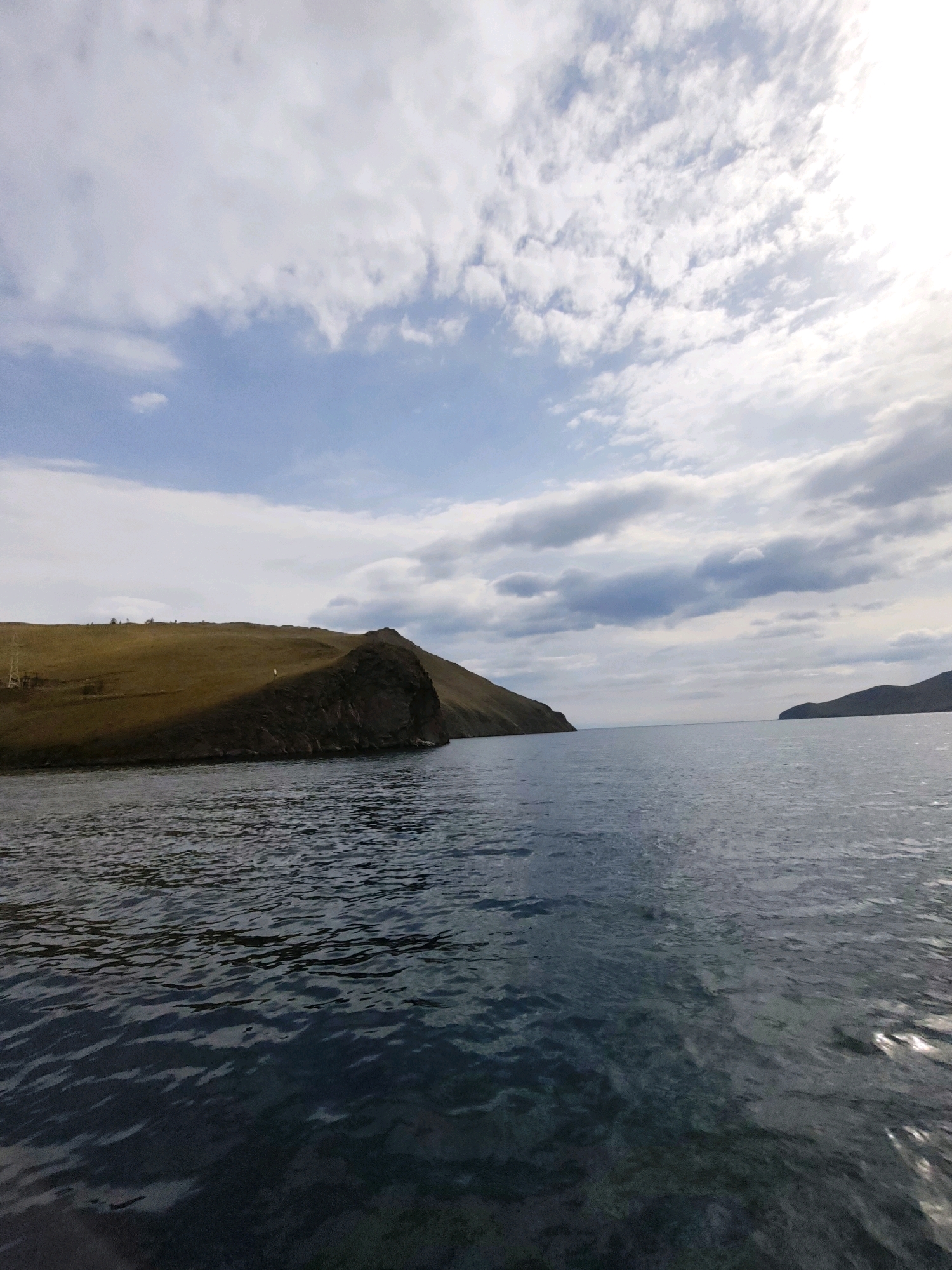 This is a wide-angle, eye-level, outdoor, landscape photograph taken from a boat on a body of water, likely Lake Baikal, with Khuzhir, Russia, as the presumed location. In the foreground, the dark blue water shows gentle ripples. On the left side of the image, a barren, grassy hill slopes down to the water's edge, with a rocky promontory jutting out into the water. The hill is cast in shadow on its western face. A thin electrical pole and wires are visible on the upper left part of the hill. In the distance, two land masses are visible on either side of a strait, with the sky above filled with a mixture of white and grey clouds, with patches of blue sky showing through. The overall lighting suggests an overcast day, possibly late afternoon or early morning. There are no people or animals visible in the frame.