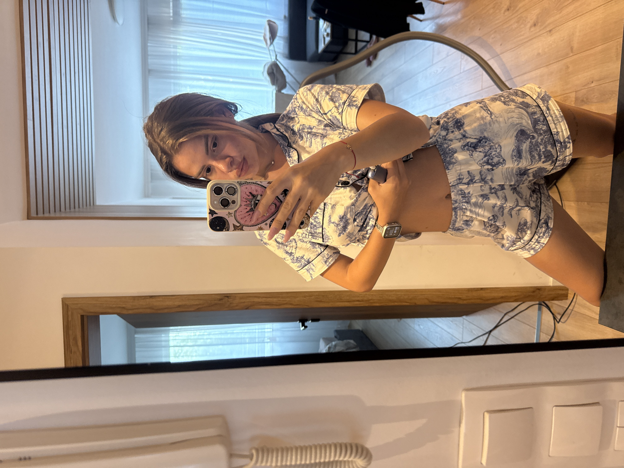 A mirror selfie captures a young woman standing in an indoor setting, likely a bedroom or hotel room. She is dressed in a matching blue and white pajama set, featuring a toile de Jouy-style pattern with pastoral scenes. She holds a smartphone, equipped with a pink, celestial-themed case, up to take the photo, slightly lifting her top to expose her midriff. Her dark hair is styled, and she has long, light-colored fingernails. On her left wrist, an Apple Watch displays "09:30" and "100%", while a red string bracelet adorns her right wrist. Her expression is neutral as she looks towards the camera lens.
The reflection in the mirror reveals a room with wooden flooring, a window with horizontal blinds, and a vacuum cleaner hose resting on the floor. In the foreground, below the mirror, are a white corded telephone and white electrical wall switches. The mirror itself has a light-colored wooden frame visible at the top.