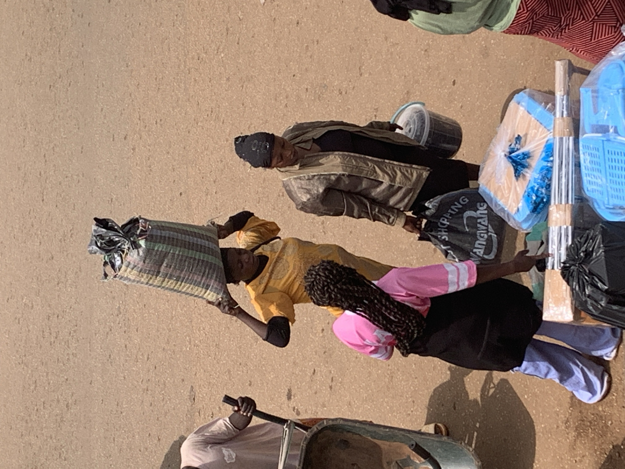 This media file captures a street scene in Bukuru, Nigeria, likely during daylight hours given the shadows. The ground appears to be a paved or dirt road, suggesting an outdoor, urban environment.
The primary subjects are several individuals, some of whom appear to be women. One woman, wearing a yellow top and black elbow-length sleeves, is carrying a large, striped bag on her head. Another woman, in the background, wears a patterned headscarf and a jacket over a black top, holding a bucket. A third person, with braided hair and wearing a pink shirt with white stripes and a black skirt, is also visible. In the foreground, a person in a light-colored shirt is pushing a cart. Several objects are scattered around, including plastic buckets, a large striped bag, and wrapped items that appear to be household goods or merchandise. Some of these items are in blue plastic bags, and one large item is covered in plastic sheeting and appears to be a broom or mop with a wooden handle. There is also a black trash bag.
The activity depicted is consistent with street vending or daily life in a busy market or roadside area where goods are transported and people are engaged in various tasks. The expressions of the individuals are not clearly discernible. The overall impression is one of a bustling, everyday scene in a Nigerian town. Some visible text on a black bag reads "SHOPPING AGWANNE".