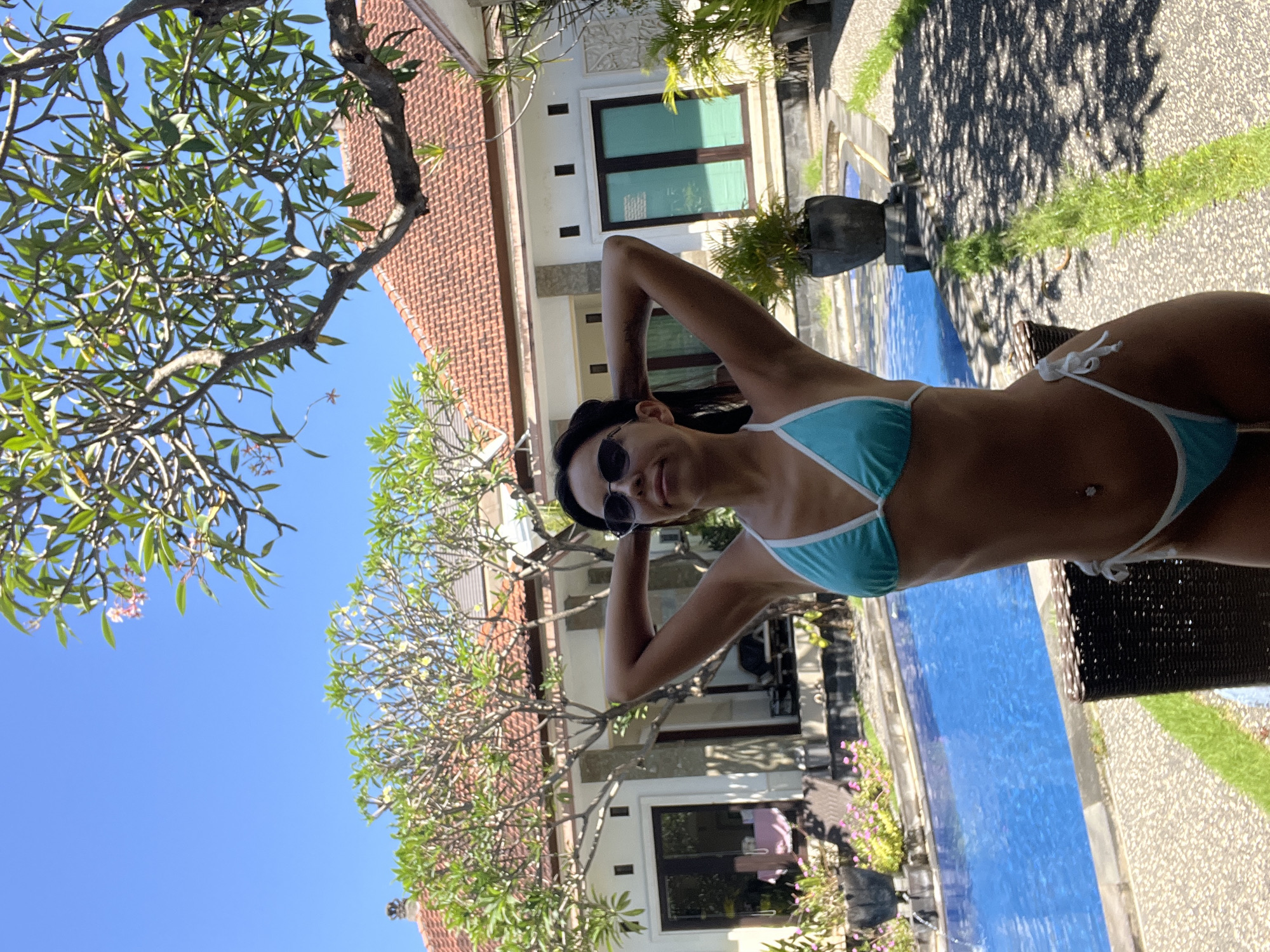 The image shows a woman in a light blue bikini sitting by a pool in Kuta Selatan, Indonesia. She has dark hair, is wearing sunglasses, and has her arms raised behind her head. The pool is long and narrow, running parallel to a building with a tiled roof and windows. Lush greenery surrounds the pool area, including trees and landscaped vegetation. The sky is clear and blue.