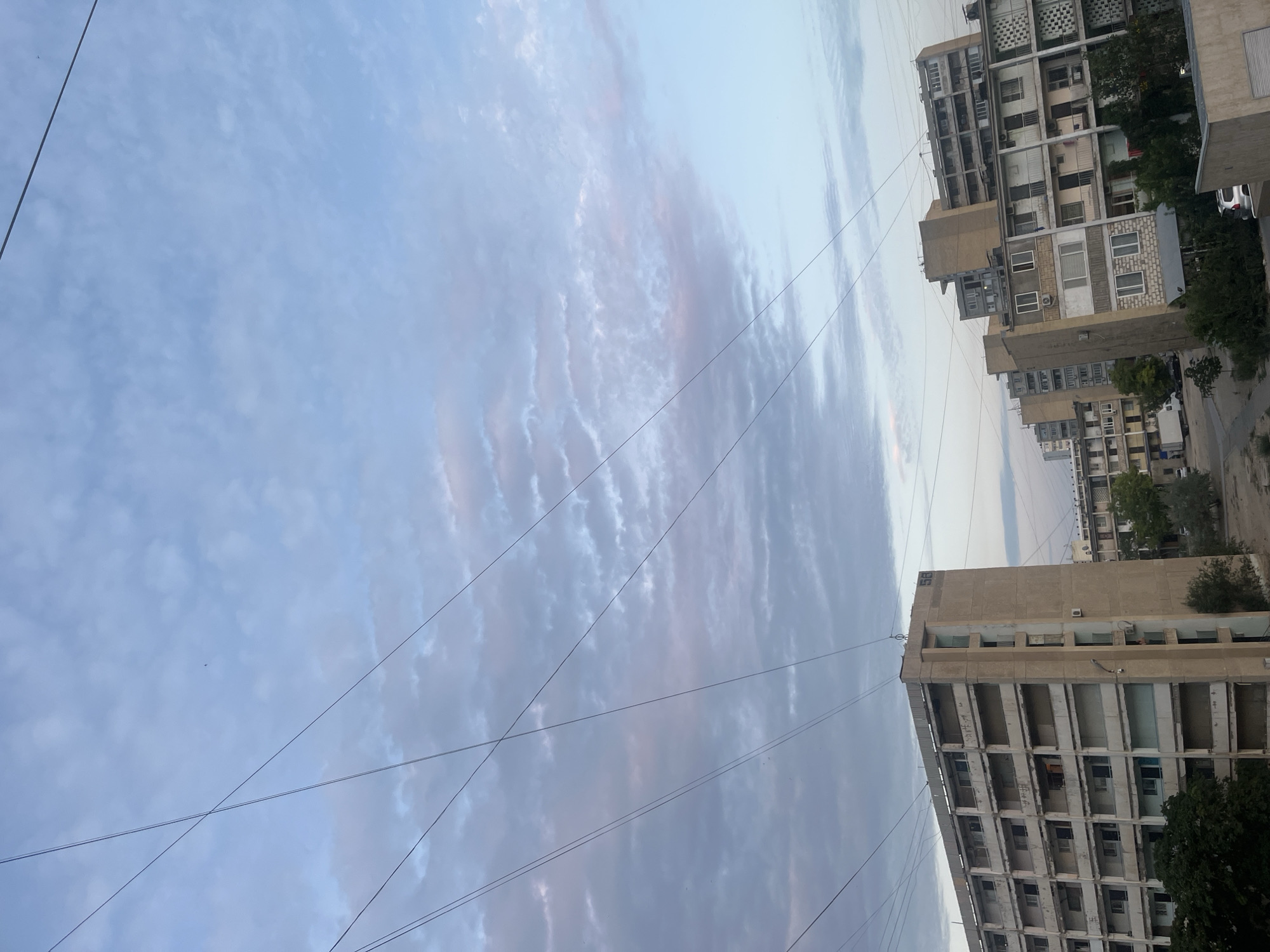 The media file shows a sky with some clouds and buildings in the background. The image is oriented vertically. The sky is a light blue with some wispy clouds, possibly indicating it is morning or evening. The clouds have hints of pink and purple, which could suggest sunset or sunrise. There are several power lines running across the sky.
In the background, there are multiple buildings, appearing to be apartment complexes. They are multi-story structures with balconies and windows. Some of the buildings look weathered and show signs of age. Trees are also visible, some of which are dark green and leafy, while others appear more sparse. The buildings are arranged in a row, suggesting an urban or suburban setting.
A car is partially visible on the right side of the image, near one of the buildings. The overall scene evokes a sense of everyday urban life, possibly in a residential area. The time of day appears to be either dawn or dusk due to the lighting and the colors in the sky. The weather seems calm, with no indication of storms or strong winds. The location is identified as Aktau, Kazakhstan. No specific activity or event is taking place; it appears to be a still shot of the environment. There is no visible text in the image.