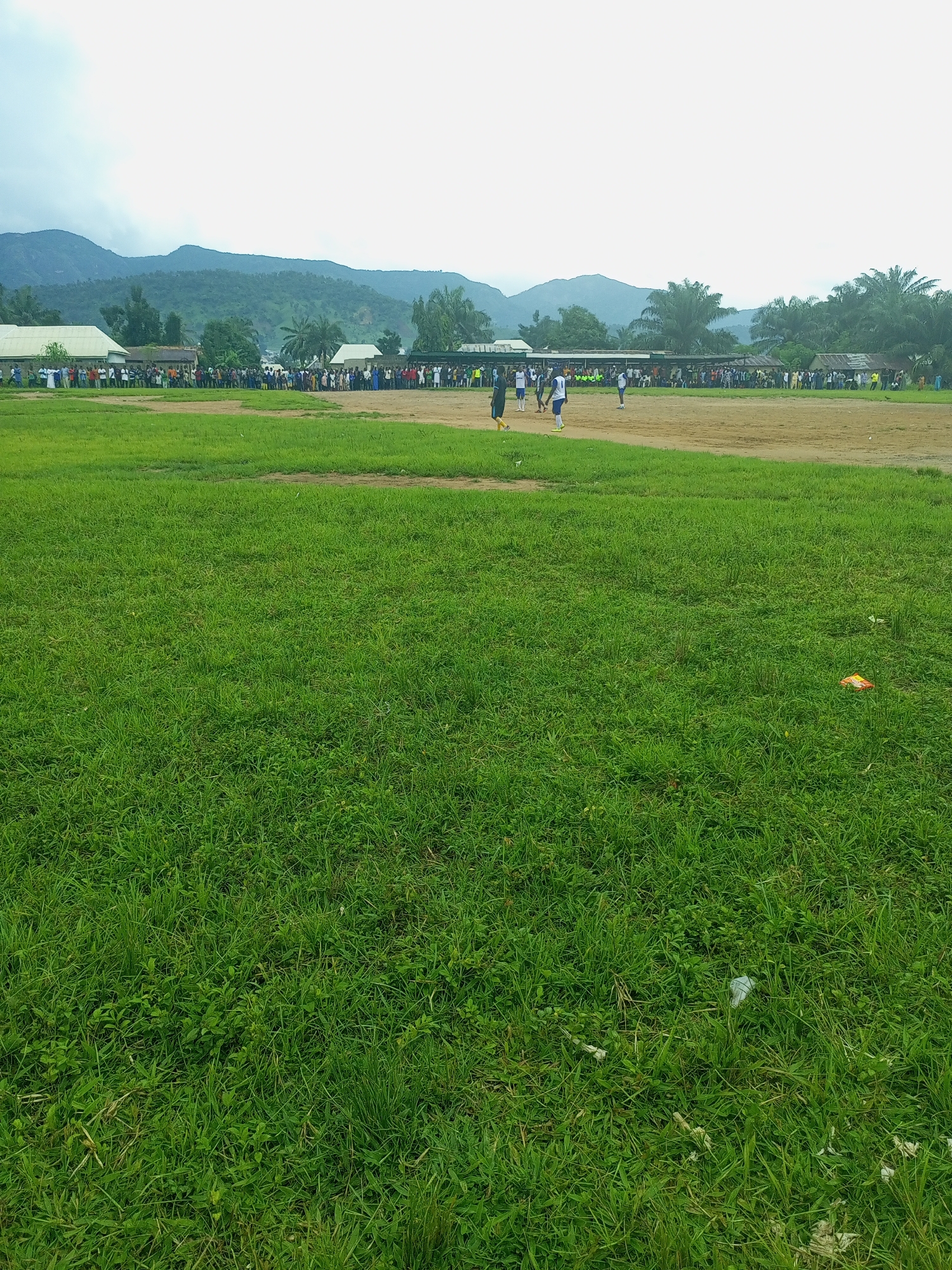 The image depicts an outdoor scene, likely a sports field, in the vicinity of Serti, Nigeria. In the foreground, a large expanse of green grass dominates the frame. In the mid-ground, a dirt field is visible with several individuals, appearing to be players or spectators, gathered. Some of these individuals are wearing athletic attire, suggesting a sporting event is in progress or has recently concluded. Behind the field, a line of buildings, possibly houses or community structures, runs horizontally. Further in the background, lush green hills and mountains are visible under a cloudy sky. The environment appears somewhat rural, with a mix of man-made structures and natural landscape.