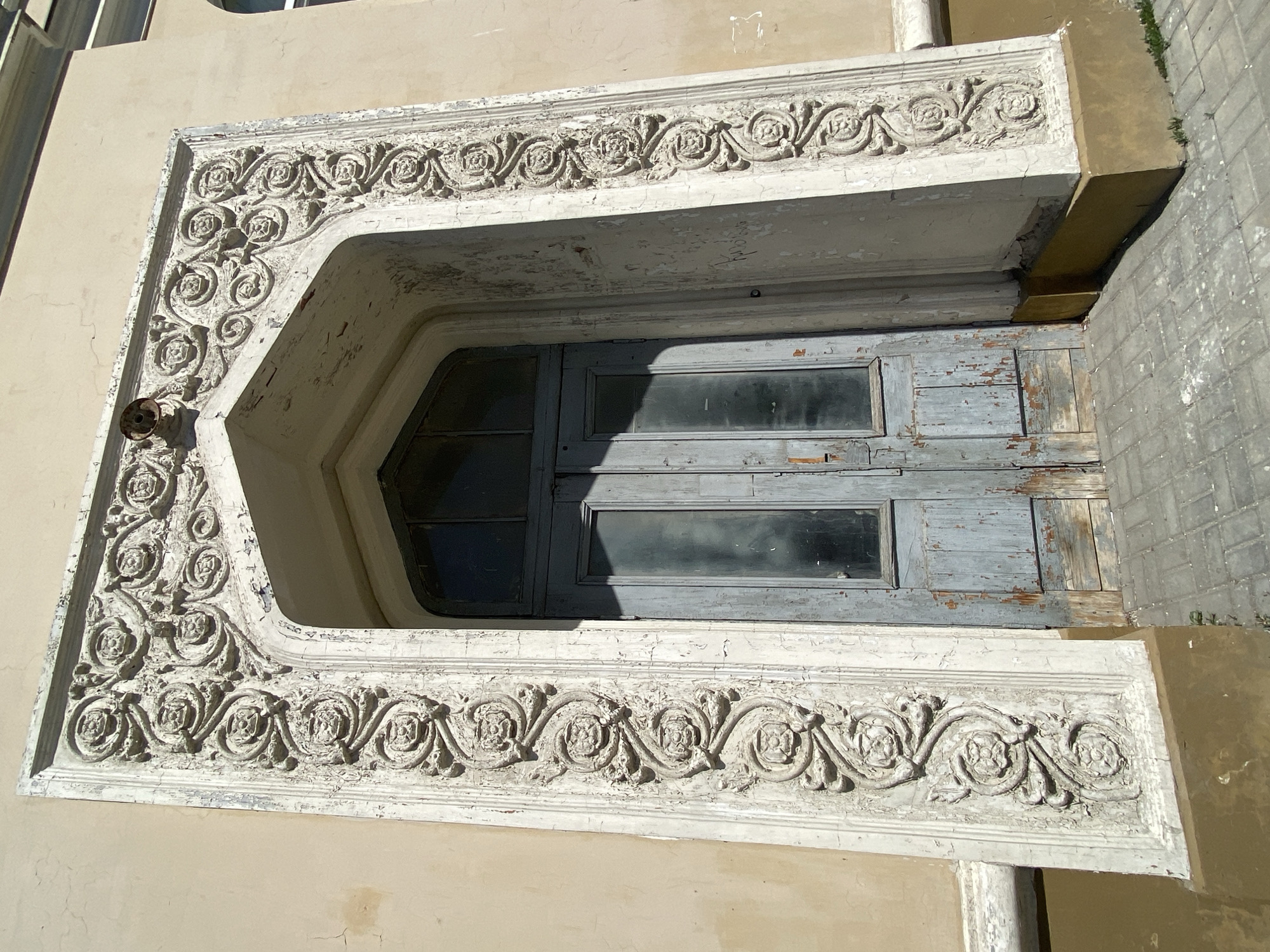 The image shows an ornate doorway, seemingly of a historical building. The doorway has a decorative, heavily textured frame with floral motifs in a repeating pattern. The frame is painted a light color, possibly white or off-white, with considerable peeling and weathering indicating age. The door itself appears to be made of wood with a series of small rectangular windows. The door has a faded blue or gray paint with similar signs of wear and tear. The building's facade is a light beige or tan color. There is a portion of a sidewalk or paved area visible to the right, and possibly parts of adjacent buildings in the background. Based on the architectural style and decorative elements, it is identified that the image depicts Kazan, Russia.