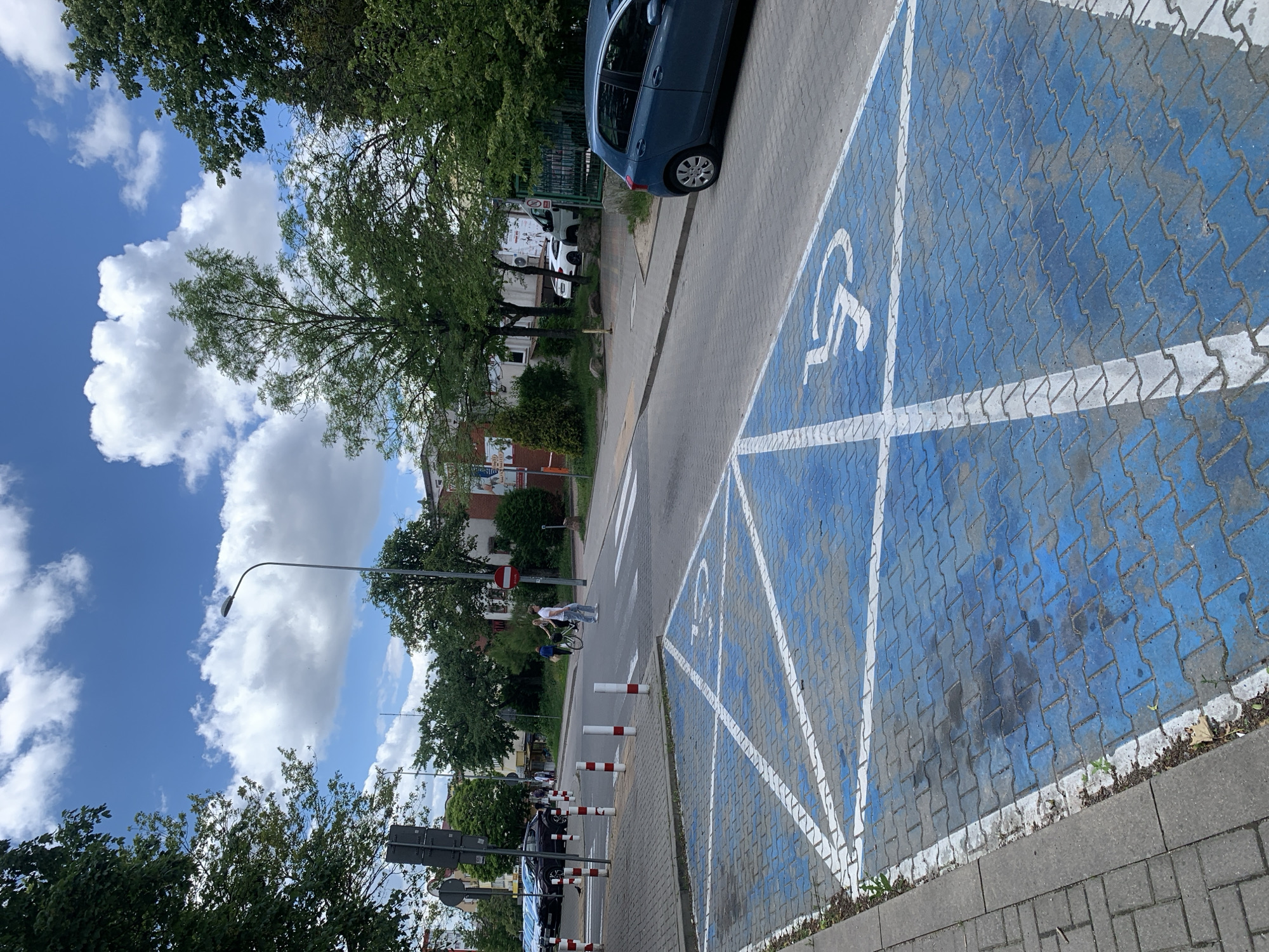 A blue-painted disabled parking spot is in the foreground of the image. White lines form a grid over the blue paint, and the International Symbol of Access is visible. The parking spot is made of interlocking gray pavers. A sidewalk runs along the left side of the parking spot. In the background, a street is visible with cars parked on the left and a pedestrian crossing. Two people are walking across the street, one on a bicycle. Trees and buildings are visible on both sides of the street. The sky is blue with white clouds. The scene appears to be in a residential or commercial area of Białystok, Poland.