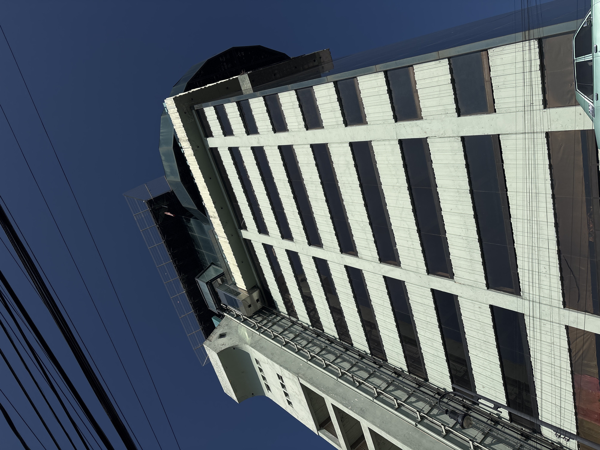 View of the City Express hotel, located in Puebla, Mexico. The perspective is from below, looking upwards at a low angle. The building is characterized by alternating white and black vertical stripes. A glass exterior elevator is visible along the building's facade. Several power lines are also present in the bottom left corner of the image. The sky is clear and blue.