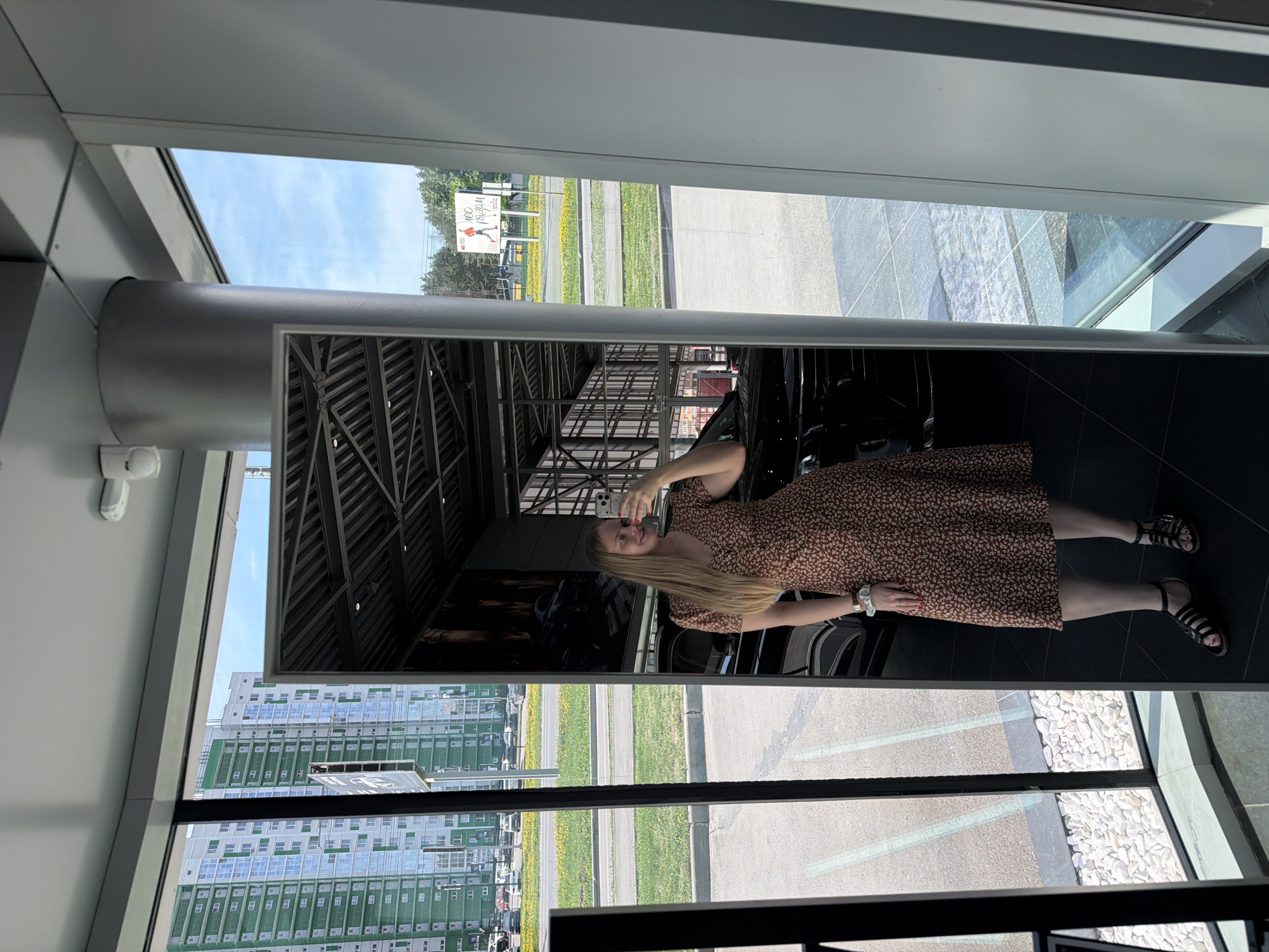 A blonde woman in a brown floral dress stands facing a mirror, holding a cell phone in her right hand, presumably taking a photo of herself. The mirror is framed in a grey metal structure, possibly inside a building with modern architecture.
Outside the window and reflected in the mirror are various views of Izhevsk, Russia. There's a glimpse of a multi-story building, likely a residential complex, with green and white colors. There is a street with manicured grass, dandelions, and a signboard. Also, part of a parking area paved with light-colored pebbles is visible.