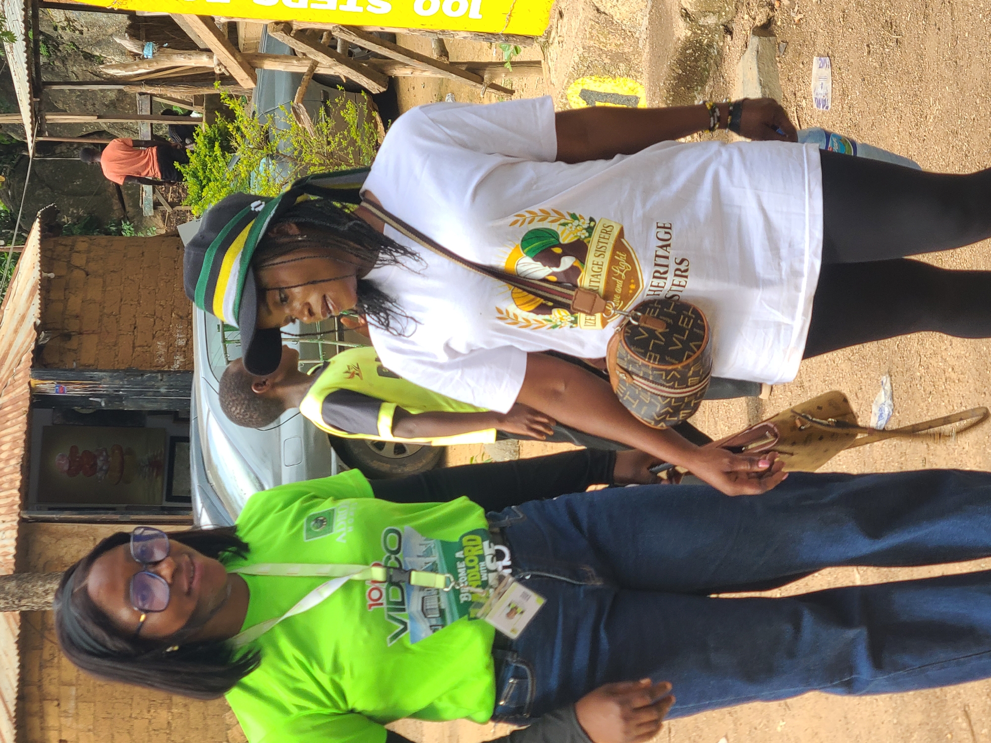 Two women are standing next to each other outside on a dirt path. The woman on the left is wearing a bright green t-shirt, blue jeans, and blue-tinted glasses. She has a lanyard with an ID card around her neck. The woman on the right is wearing a white t-shirt with a circular graphic on it, black leggings, and a black hat with green, yellow, and white stripes. She is carrying a brown purse with a logo pattern on her left shoulder and holding a small brown purse in her right hand. A young boy in a yellow and grey t-shirt is partially visible behind the woman on the left. In the background, there are buildings with corrugated metal roofs and a vehicle. A yellow sign with black text is visible on the right side of the image. The setting appears to be an outdoor market or street in Jos, Nigeria.