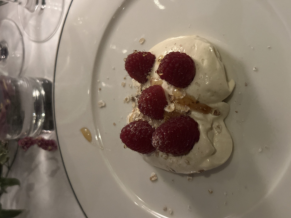 A close-up captures a dessert plate featuring a dollop of creamy white substance adorned with five raspberries, glistening with a light glaze or syrup. The base appears to have a sprinkling of oats or granola. The plate is white with a thin, dark green line along the rim. A partial view of glasses and a floral arrangement sit adjacent to the plate, suggesting a dining setting.