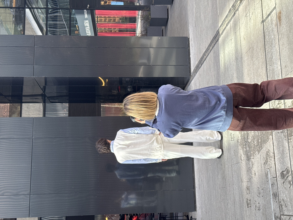 The image displays an outdoor scene at the Galleria Tbilisi shopping mall in Tbilisi, Georgia. The location is specifically near Askaneli Restaurant, as indicated by a reversed reflection of "ASKANELI RESTAURANT" visible on the building's reflective surface. In the immediate foreground, a person with short, light brown hair, wearing a dark blue long-sleeved top and dark reddish-brown wide-leg pants, stands with their back to the camera. This individual is holding a black smartphone with both hands, aimed forward as if taking a picture or video. Directly in front of this person, another individual stands with their back to the camera, positioned against a dark, highly reflective building facade. This second person has dark brown hair and is dressed in a light blue collared shirt, a cream-colored sleeveless vest, and white wide-leg trousers, with black shoes visible. A blurred reflection of a figure, possibly the photographer, is visible on the dark wall to the left of the posing individual. The building itself features large, dark, multi-panel reflective surfaces. Reflections on the right side of the building show an escalator and red-curtained interior elements. The ground is paved with light-colored square tiles, and dark, rectangular planters containing minimal vegetation are visible on the right. The scene depicts a photographic interaction between two individuals in an urban environment.