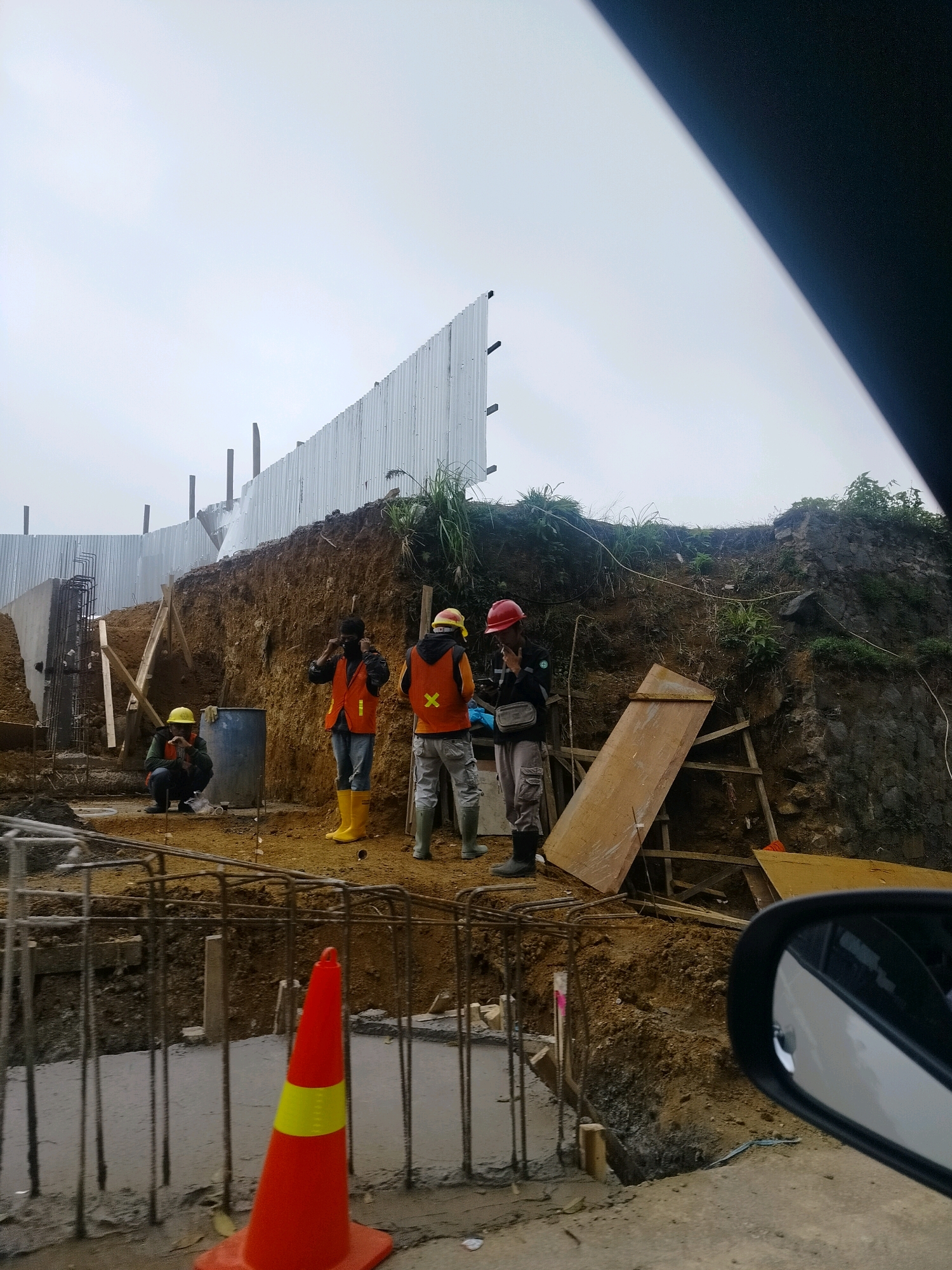The image depicts a construction site on a hillside, possibly in Cisarua, Indonesia, given the context. A corrugated metal fence runs along the top of the elevated area, separating it from the sky. The ground is composed of loose soil and rocks, with several workers present.
In the foreground, there is rebar and concrete work. A bright orange traffic cone with a yellow reflective band is prominently placed. To the left, a construction worker wearing a yellow hard hat, an orange safety vest, and dark pants is squatting near a blue barrel.
Further back, three construction workers are visible. One, wearing a yellow hard hat and orange vest, is standing and facing away from the camera. Another, to his left, is wearing a red hard hat, an orange vest, and appears to be adjusting something on his face, possibly a mask. A third worker, also in an orange vest and red hard hat, is standing and facing the camera, smoking a cigarette and holding a small bag.
The environment appears to be outdoors with an overcast sky. The hillside is uneven and shows signs of excavation. Various construction materials, including wooden planks and scaffolding, are scattered around the site. A car's side mirror is visible in the lower right corner of the frame, indicating the photograph was taken from inside a vehicle.