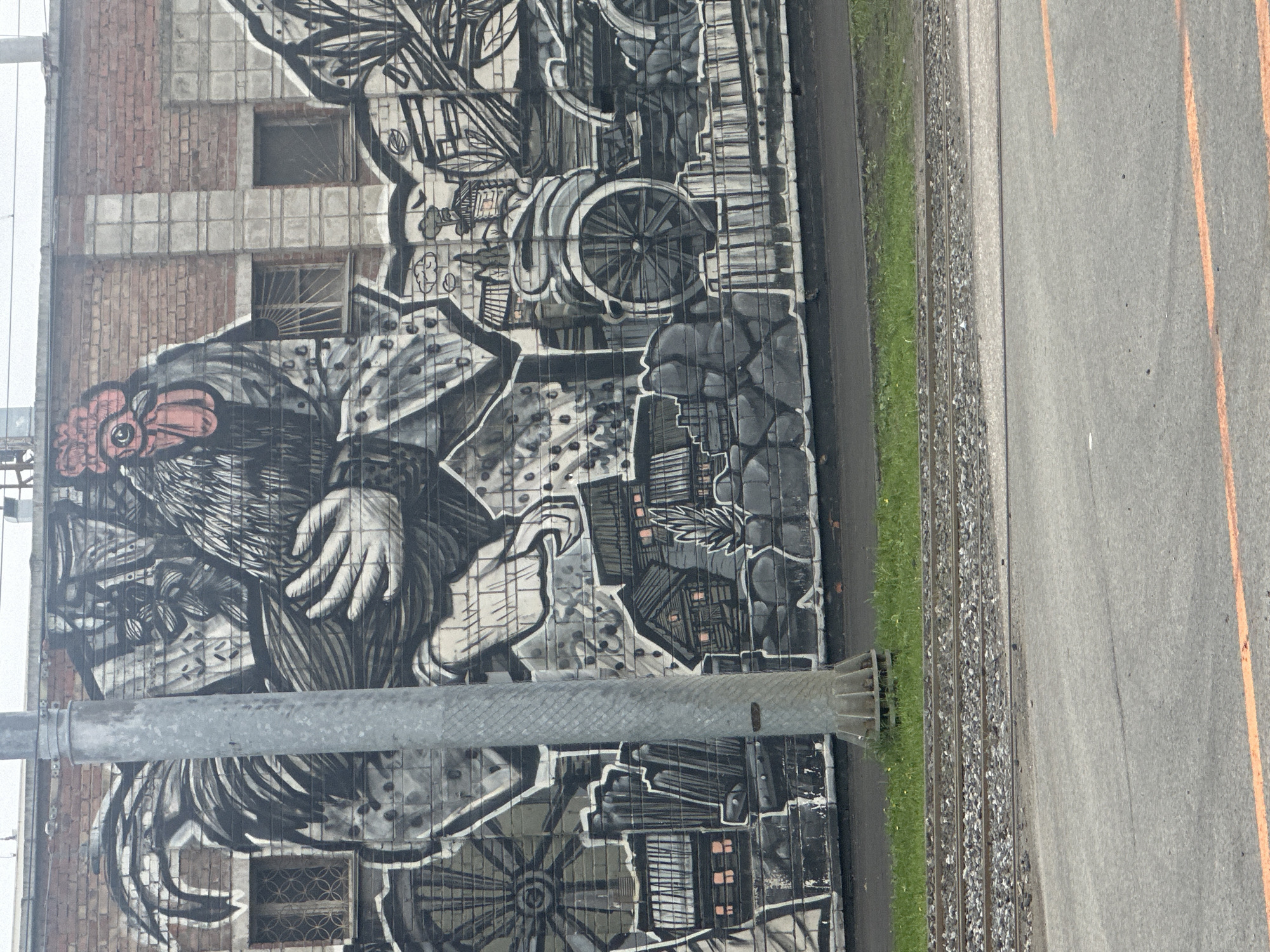 A mural covers a brick wall, featuring a monochromatic, stylized rendering of a rooster and various city elements. The mural is beside a road with yellow lane markers and a patch of grass next to train tracks. A metal pole stands in front of the mural. The style is graffiti, with complex linework depicting architecture, vehicles, and other urban imagery. The scene is near train tracks, suggesting the industrial district of Novosibirsk, Russia, known for street art.