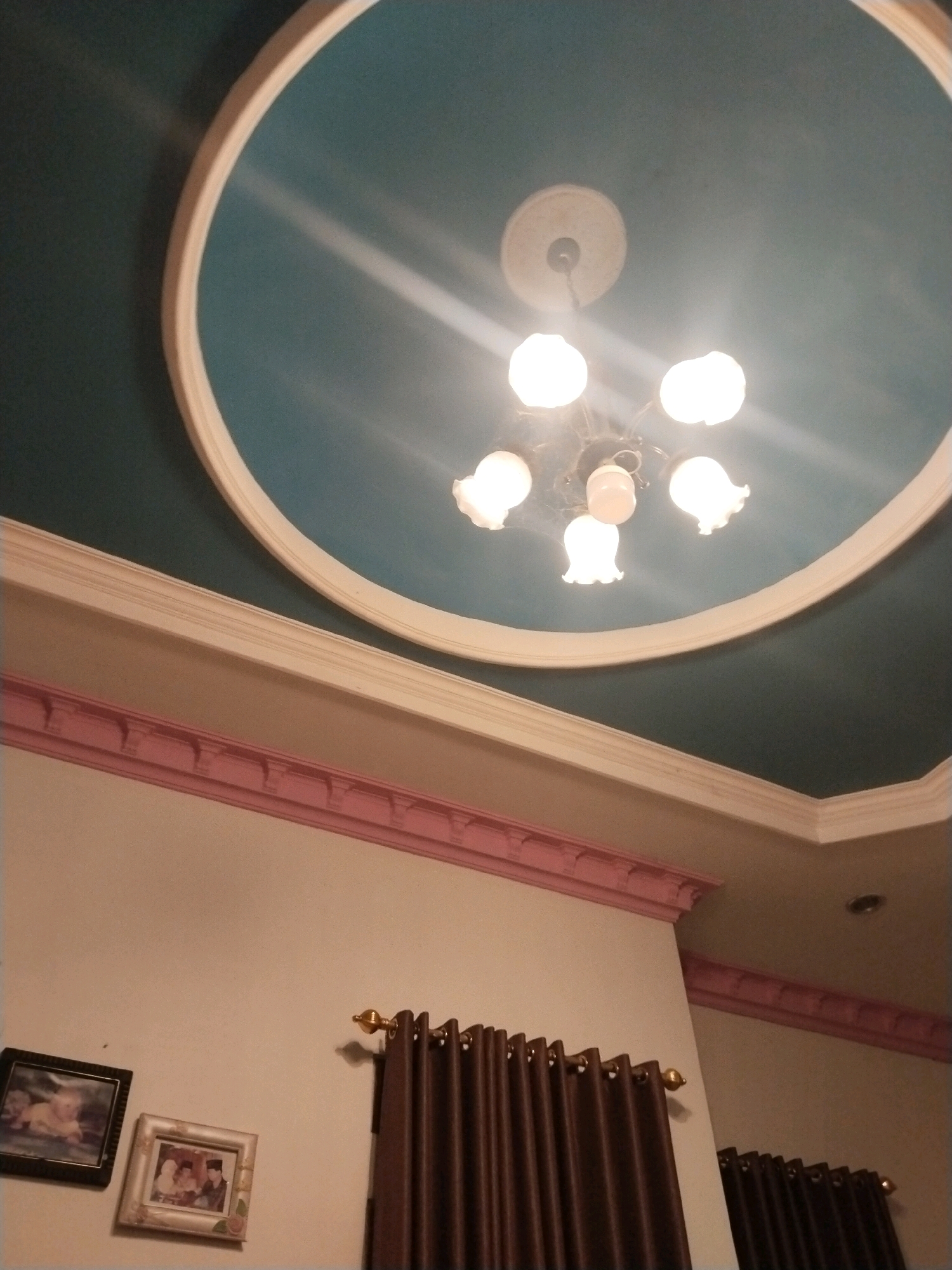 This is a photograph taken inside a room, likely a residential interior, located in Jembayat, Indonesia. The ceiling is painted a teal color and features a circular white border. A chandelier with five light bulbs hangs from the center of the ceiling, illuminating the space. Below the ceiling, decorative molding painted pink runs along the top of the walls. On the wall, two framed photographs are visible. The photograph on the left is framed in black and appears to show a baby. The photograph on the right is framed in a light, ornate pattern and shows what looks like a group of people. To the right of the photographs, a pair of dark brown curtains hangs from a gold curtain rod. Another set of similar curtains is visible further to the right. The overall lighting suggests the photo was taken in the evening or in a dimly lit environment, relying on the chandelier for primary illumination.