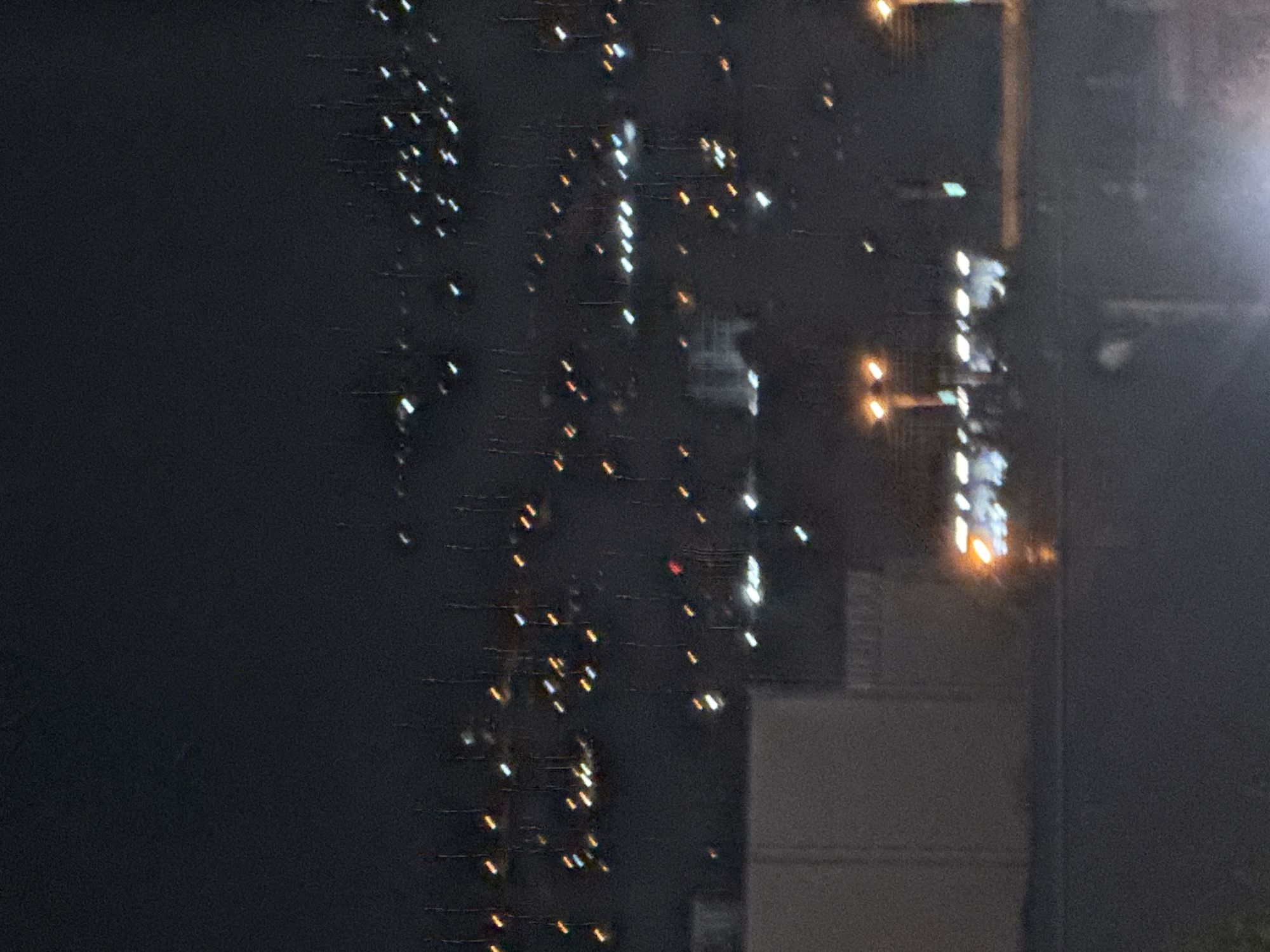 The image depicts a nighttime scene in Tbilisi, Georgia, with a blurred view of city lights and structures. The foreground features a large, light-colored building on the left and a darker, indistinct area that might be a road or open space. In the mid-ground, a line of illuminated streetlights is visible, casting a warm glow. Further in the background, the cityscape is characterized by scattered points of light, suggesting buildings and urban development on a slope. The overall impression is one of a city at night, viewed from a distance, with a slightly grainy quality to the image due to low light conditions.