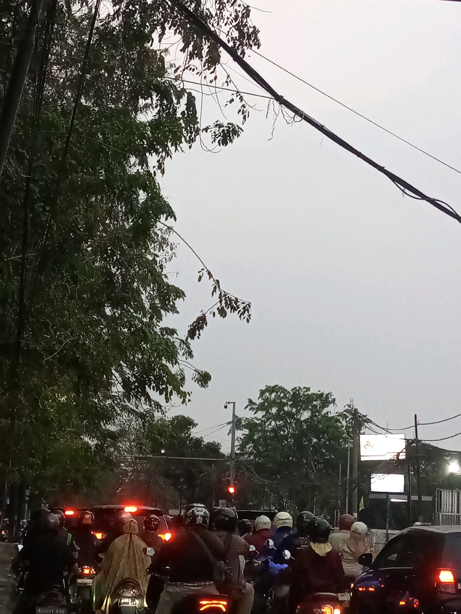 A traffic jam is depicted on a street in Bekasi, Indonesia, under a cloudy sky. A line of motorcycles and cars are stopped at what appears to be a traffic signal, which is illuminated red. Several individuals are riding motorcycles, many wearing helmets. Some motorcycles are equipped with rear boxes or bags. Trees line the street, and overhead wires are visible. In the background, buildings and streetlights can be seen. The overall scene suggests a common urban traffic situation in the region.