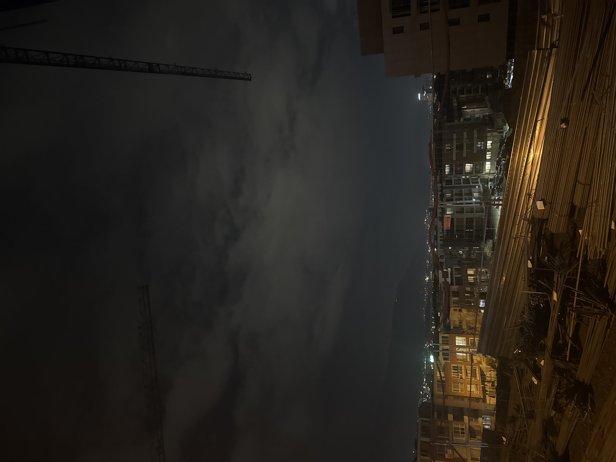 At night, a cityscape is visible from an elevated position. To the right, a structure appears to be under construction, with building materials stacked and rebar visible. In the background, lit buildings suggest an urban residential area. Two construction cranes are noticeable against a dark sky covered by patchy clouds. Based on the architecture and urban layout, the location is in Sofia, Bulgaria. There are no people visible in the image.