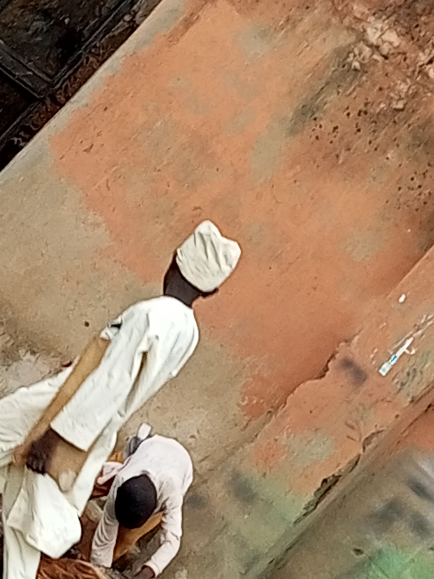 The image shows two individuals in Gombe, Nigeria, on a dusty, reddish-brown, uneven surface that appears to be a pathway or ground near a structure. In the foreground, a person is bent over, seemingly engaged in an activity on the ground. This individual is wearing a white shirt with long sleeves and dark pants. Behind this person, a second individual stands, facing away from the camera. This person is dressed in a white robe and a white head covering. The background consists of the same textured, reddish-brown surface with some darker, possibly metal, elements visible in the upper left corner. The overall scene depicts a casual, outdoor setting within the city of Gombe.