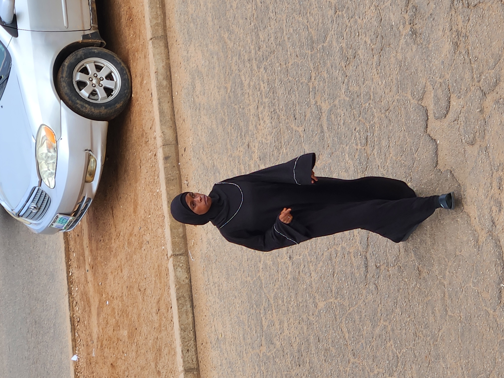 A woman walks on a cracked asphalt road in front of a beige curb and a dirt shoulder. She is wearing a black abaya and hijab, with decorative white trim on the sleeves and neckline. A silver car is parked on the dirt shoulder. The car has a license plate that reads "KUL 558X". The woman is looking to her right and appears to be walking towards the viewer. The scene is outdoors, under natural light.