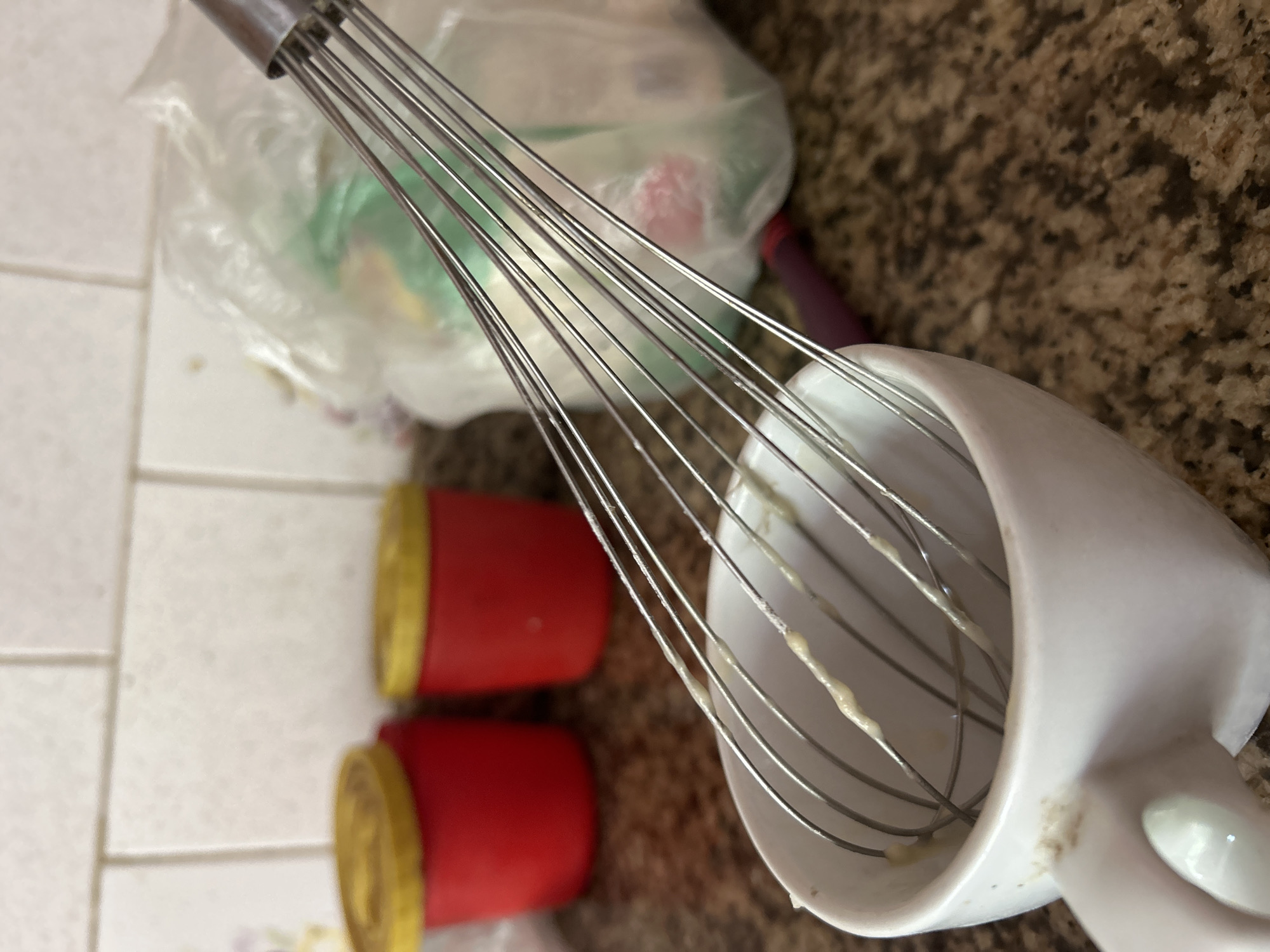 The image depicts a close-up view of a white whisk inside a white ceramic mug, with some yellowish residue clinging to the whisk wires and the inside rim of the mug. In the background, two red cylindrical containers with yellow lids are visible, along with a translucent plastic bag containing unseen items, possibly food ingredients. The surface beneath the objects appears to be a speckled granite countertop. The scene is set against a backdrop of white tiled wall. The composition suggests a domestic kitchen environment. The photograph was taken in Santa Ana, Venezuela.