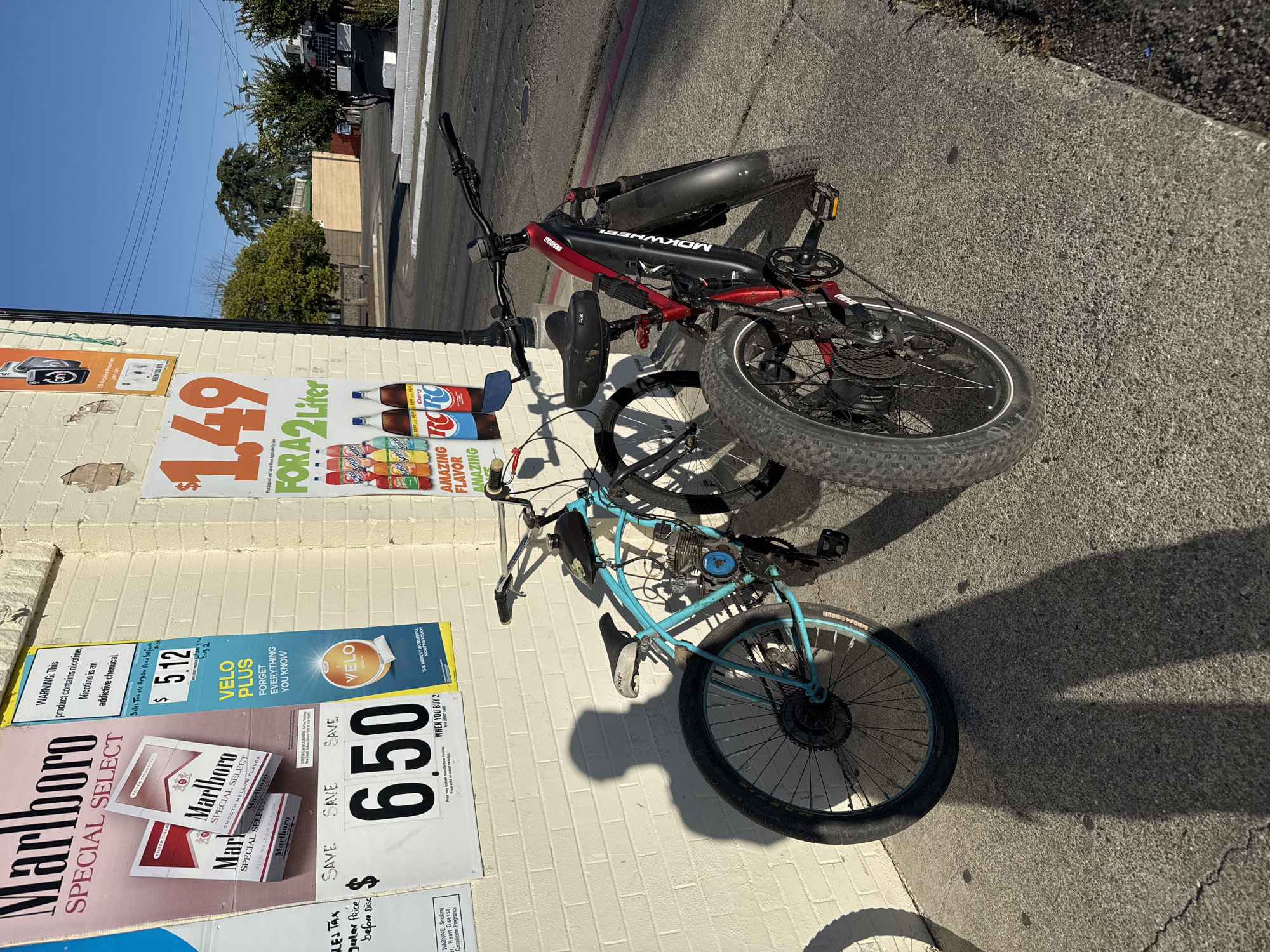 The image displays an outdoor scene in what appears to be an urban or suburban setting, possibly in Rio Linda, United States. Two bicycles are leaning against a light-colored brick building on a sunny day, indicated by the bright blue sky and distinct, elongated shadows suggesting either early morning or late afternoon.
One bicycle is a red and black electric bike, with "MOKWHEEL OBSIDIAN" visible on its frame, featuring robust, knobby tires. Leaning in front of it is a light blue bicycle that has been customized with a small engine kit, including a motor mounted near the pedals and a fuel tank on the top tube, making it a motorized bicycle. Both are parked on an asphalt surface next to the building.
The building's wall is covered with several advertising posters. One poster advertises RC Cola and other sodas for "$1.49 FOR A 2 Liter," mentioning "AMAZING FLAVOR" and "Cherry." Below this, a Marlboro Special Select cigarette advertisement promotes a "SAVE 650 WHEN YOU BUY 2" deal and includes a health warning. Another sign advertises "VELO PLUS" nicotine pouches, offering to "SAVE $5.12 WHEN YOU BUY 2" and carrying a warning about nicotine being an addictive chemical. In the background, trees, power lines, and distant structures are visible under the clear sky. No people are present in the image.