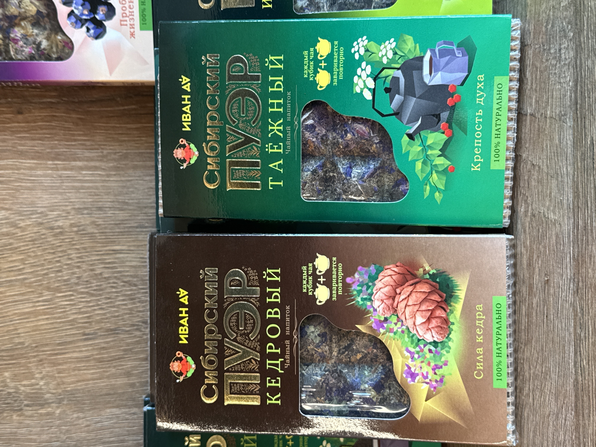 The image displays two cardboard boxes of herbal tea products. The box on the left is brown and features gold lettering that reads "ИВАН ДУ" and "Сибирский ПУЭР КЕДРОВЫЙ" with an illustration of pine cones and flowers. The box on the right is green and has gold lettering that reads "ИВАН ДУ" and "Сибирский ПУЭР ТАЁЖНЫЙ" with an illustration of a teapot, a mug, and berries. Both boxes have a window revealing the tea leaves inside and a green banner at the bottom stating "100% НАТУРАЛЬНО" (100% Natural). The boxes are placed on a wooden surface, likely a shelf. The background shows a textured wooden wall. The lighting appears to be natural, casting a soft glow on the products. The image does not provide information about a city in Russia or specific locations within it.
