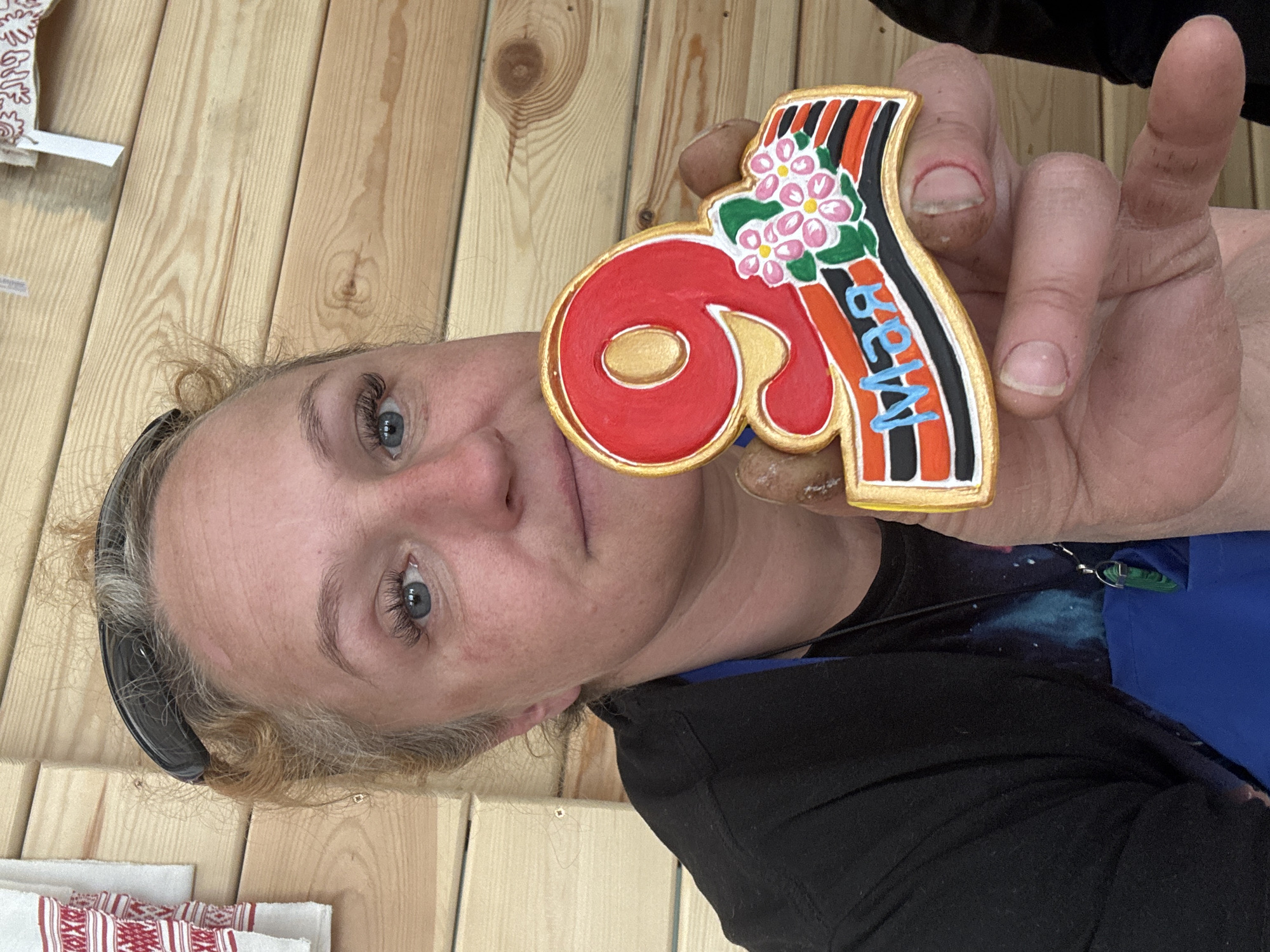 A woman is holding a decorative souvenir that is likely from Chaikovsky, Russia. The souvenir is shaped like the number 9 with the word "May" in Russian written on it. The souvenir has a gold outline and is decorated with a cluster of pink flowers and green leaves. The woman has blue eyes, fair skin, and light blonde hair. She is wearing a black shirt with a galaxy print and a blue apron. The background consists of a wooden wall and other textiles.