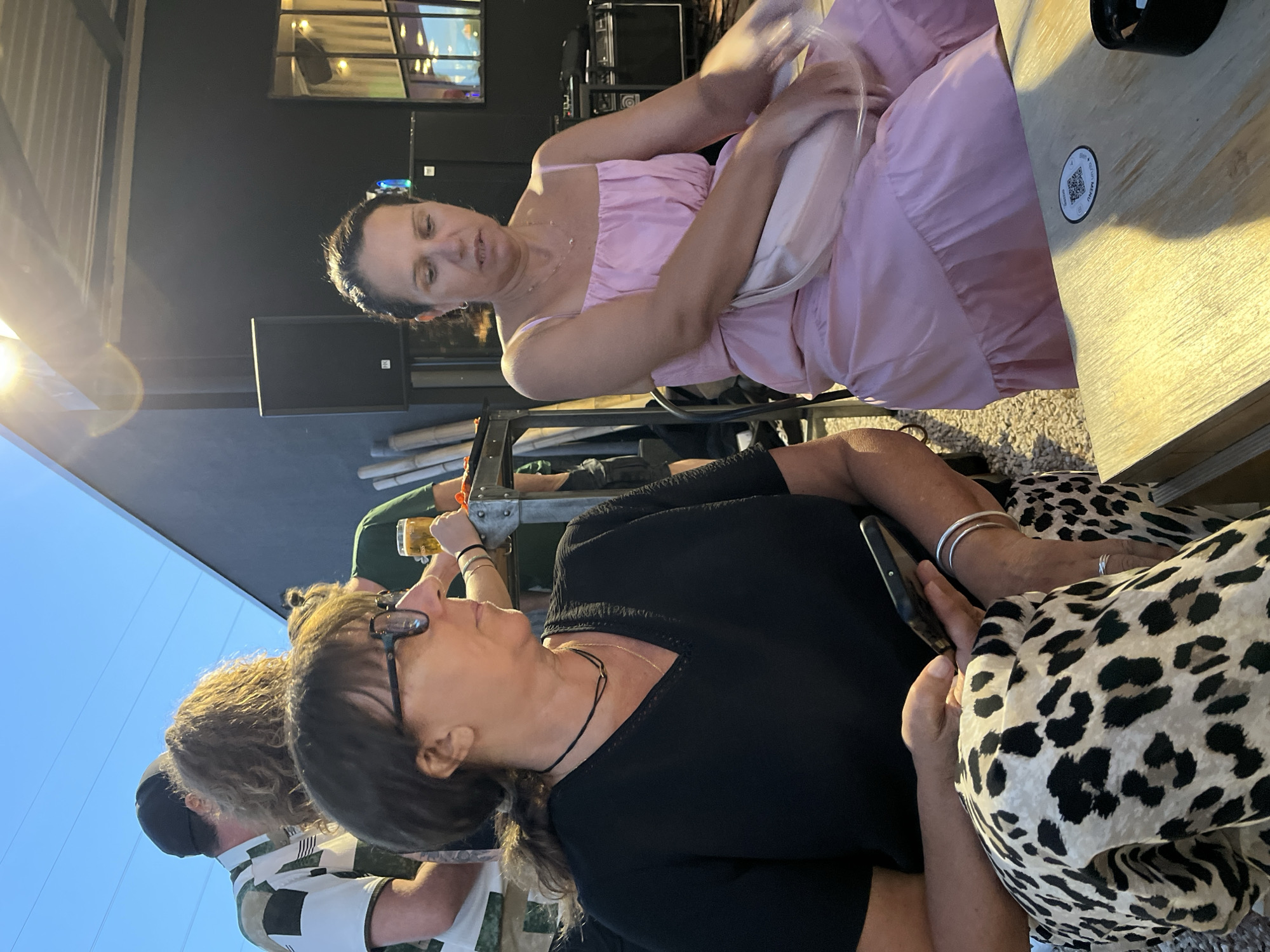 In Savigneux, France, two women are seated at a table outdoors. The woman on the left, with glasses and brown hair tied back, wears a black top and a leopard-print skirt. She is holding a smartphone. The woman on the right, with dark hair, wears a light pink dress and is looking down. A light-colored purse rests on her lap. Behind them, other individuals and outdoor furniture are visible. The setting appears to be a restaurant or bar with a dark-colored building and a canopy overhead. The lighting suggests it is late afternoon or early evening.