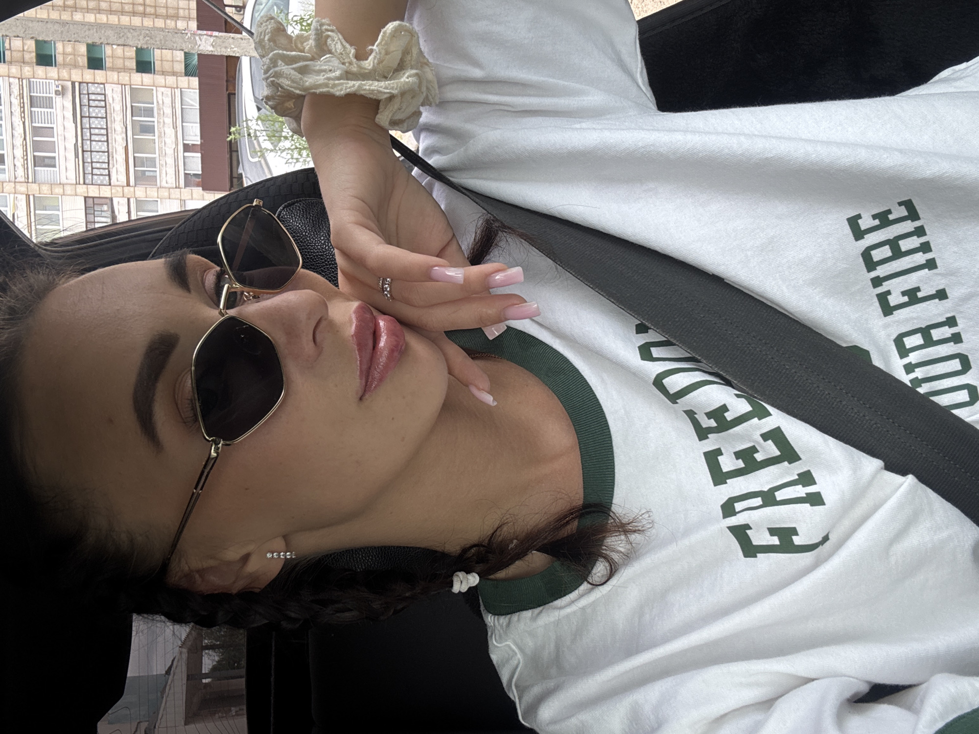A woman with dark braided hair and sunglasses is seated in a car, her right hand resting on her chin. She is wearing a white t-shirt with green lettering that reads "FREEDOM" and "YOUR FIRE". A black seatbelt is visible across her chest. In the background, a multi-story building and a concrete pillar are visible. The scene appears to be in an urban environment, possibly a residential area in Chaikovsky, Russia, given the architectural style of the building.