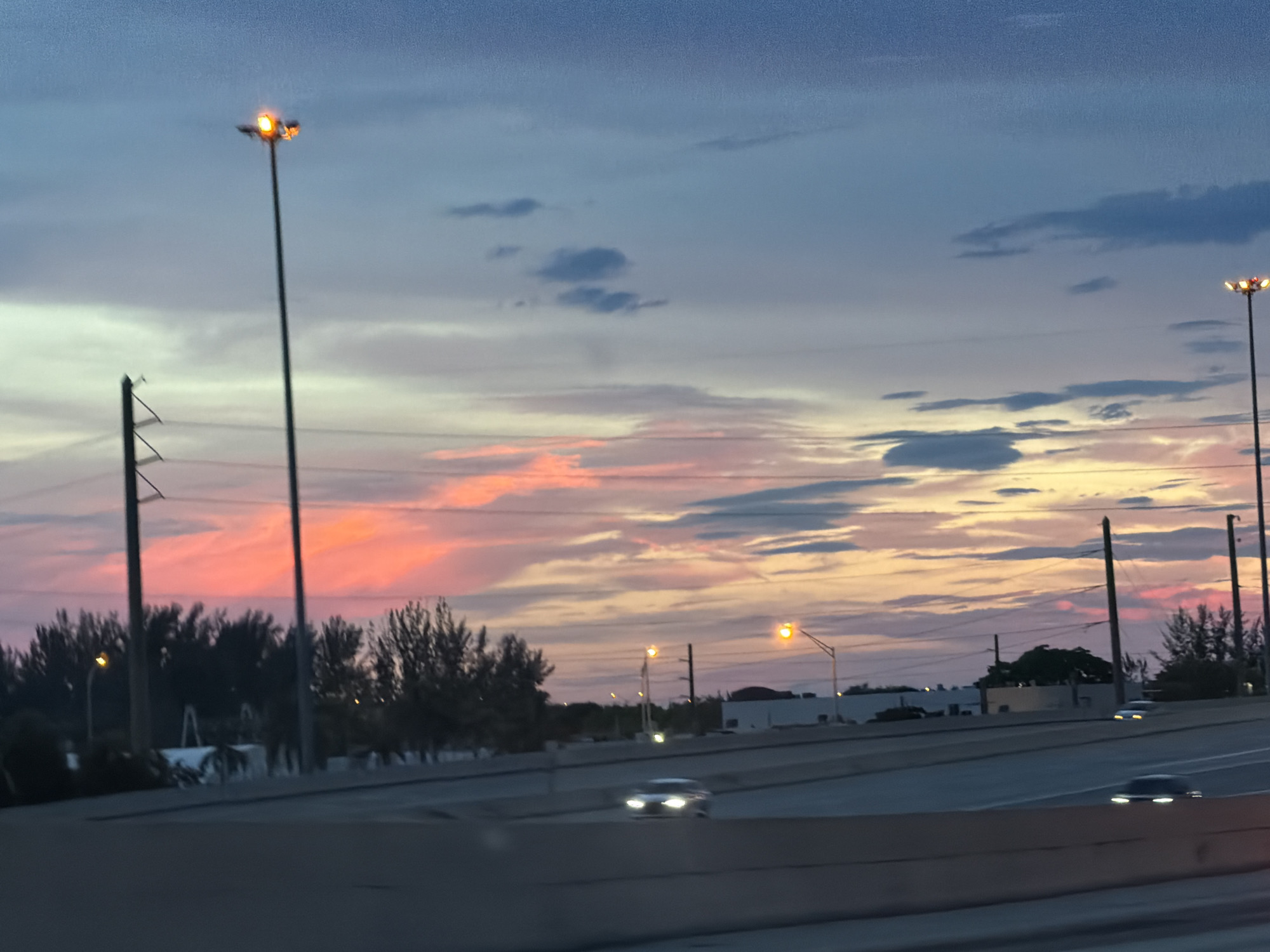 This is an eye-level, outdoor, medium shot of a highway at dusk in Miami, United States. The sky is filled with hues of pink, orange, and blue from the sunset, with scattered clouds. A tall light post with orange lights is visible on the left side of the image. There are several cars on the highway, with their headlights on, and trees line the side of the road in the background. The image is slightly blurred, suggesting it was taken from a moving vehicle.