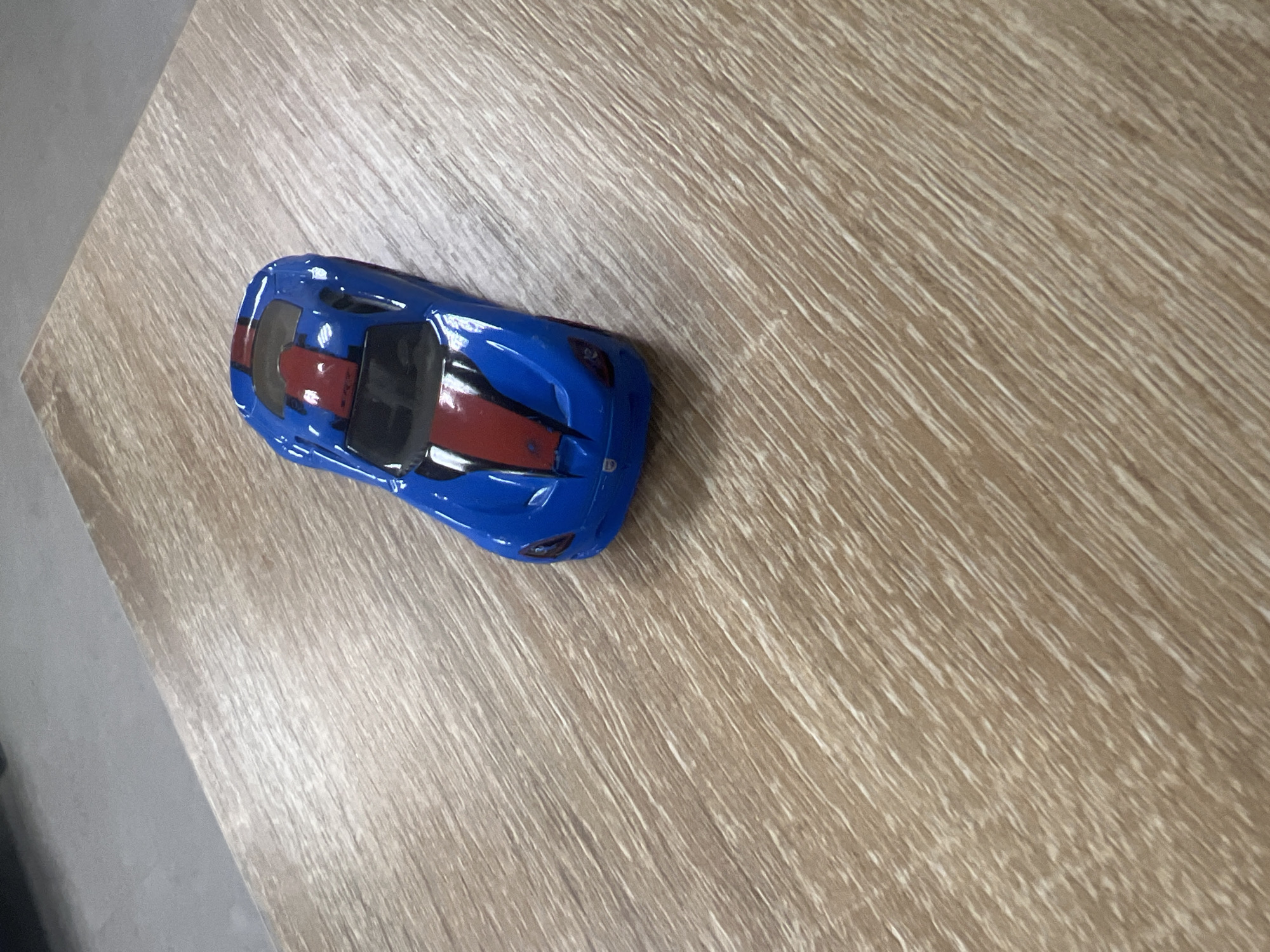 A blue toy sports car with red racing stripes sits on a wooden surface. The surface has a light brown wood grain pattern. The car is positioned at an angle, facing towards the upper right corner of the image. The background is out of focus and appears to be a grey, flat surface. The lighting suggests an indoor setting.