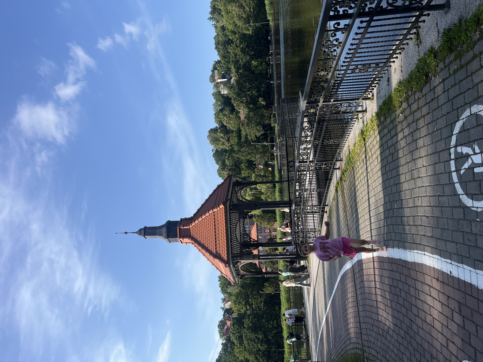 In Svetlogorsk, Russia, a young girl with brown hair, wearing a pink t-shirt and shorts, runs towards the camera on a paved walkway lined with red and grey bricks. The walkway curves to the left, and a white line marks a path on the ground, featuring a painted symbol that appears to be a bicycle. In the background, a decorative gazebo with a red tiled roof stands on a bridge or elevated walkway. Beyond the gazebo, a tranquil lake is visible, bordered by a black metal railing. The distant shore of the lake is covered with lush green trees, and several buildings, some with red roofs, are nestled among the foliage. The sky is a clear blue with scattered white clouds. Several other people are walking and standing on the walkway and near the gazebo, appearing to enjoy the scenery.