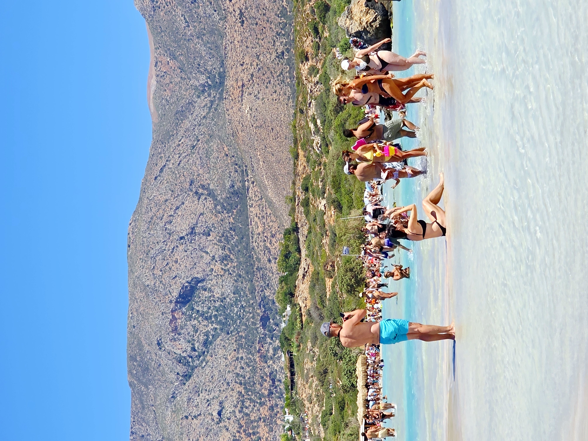 This is a wide shot photograph of a crowded beach with crystal clear turquoise water in the foreground. A man in blue swim trunks and a grey baseball cap is standing in the shallow water, facing away from the camera and holding a cell phone. Behind him, a large group of people are in the water and on the beach. Some are wearing swimsuits, while others are dressed in casual beachwear. In the background, there are green hills and mountains under a clear blue sky. A Greek flag is visible on the beach. The scene depicts a lively summer day at a popular tourist destination in Greece.