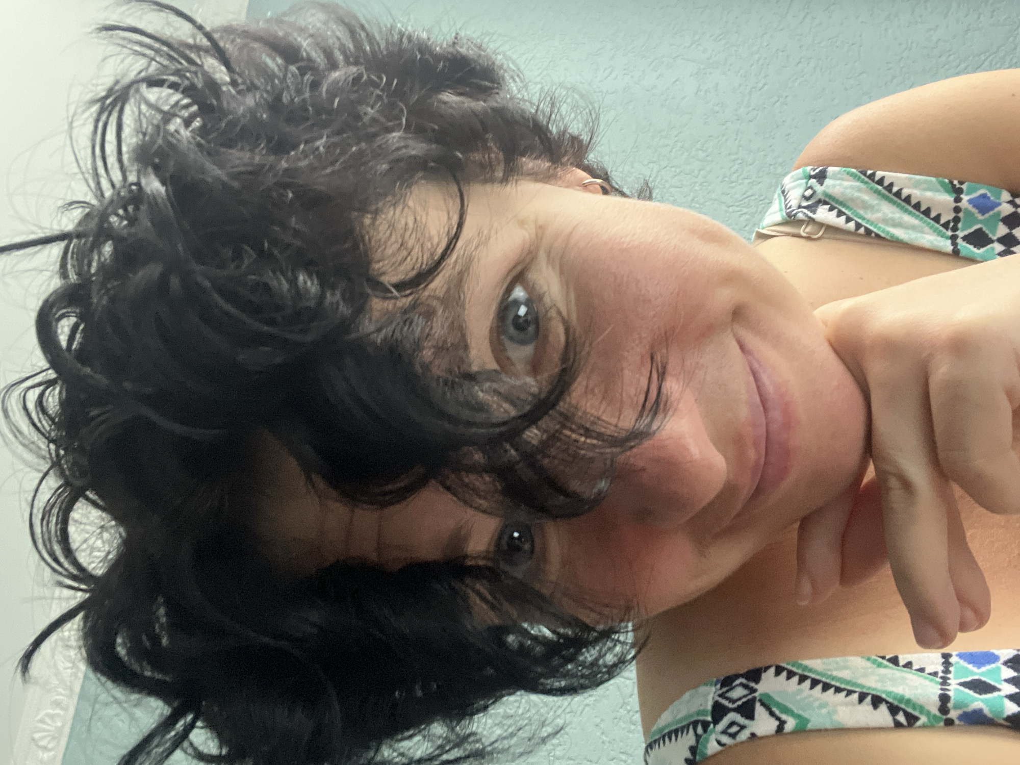 A close-up, chest-up selfie shows a woman with dark, curly, tousled hair framing her face. She has blue eyes and a light complexion with some freckles. Her left hand is resting under her chin, and she is wearing a sleeveless top with a geometric pattern of white, teal, and black. Her expression is soft, with a slight smile. The background is a light blue textured wall. The image was taken in the city of Tbilisi, Georgia.