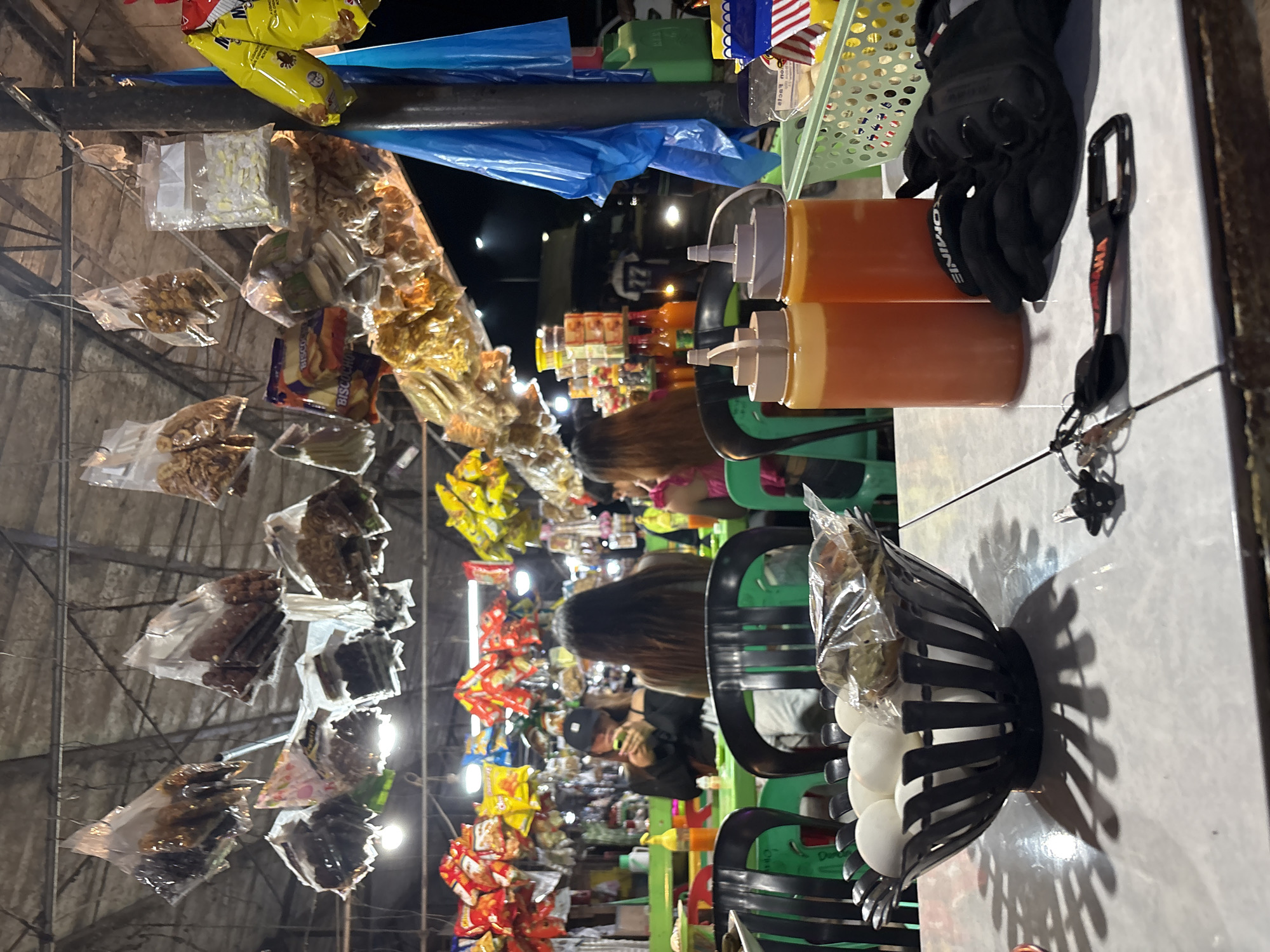The image depicts an outdoor food stall scene at night in Carmen, Philippines. In the foreground, a black, decorative metal bowl holds several white eggs, partially covered by a plastic bag containing snacks. To the right of the bowl are two squeeze bottles filled with an orange liquid, likely condiments. A pair of black gloves and a keychain with a black and red lanyard lie on the table in front of the bottles. The table's surface is white and tiled, with a prominent shadow cast by the bowl of eggs.
In the midground, several people are seated at a green table. One individual, wearing a black baseball cap and black shirt, is looking at their phone. Another person with long brown hair is visible from behind. Green plastic chairs surround the table.
The background reveals the bustling environment of the food stall. Various packaged snacks and goods are hanging from the ceiling or displayed on shelves. Bright lights illuminate the area. A blue tarp is visible, partially covering a pole. In the distance, other vehicles and structures can be seen, suggesting a busy street or marketplace setting. The overall impression is one of a lively, casual dining experience at a roadside eatery.