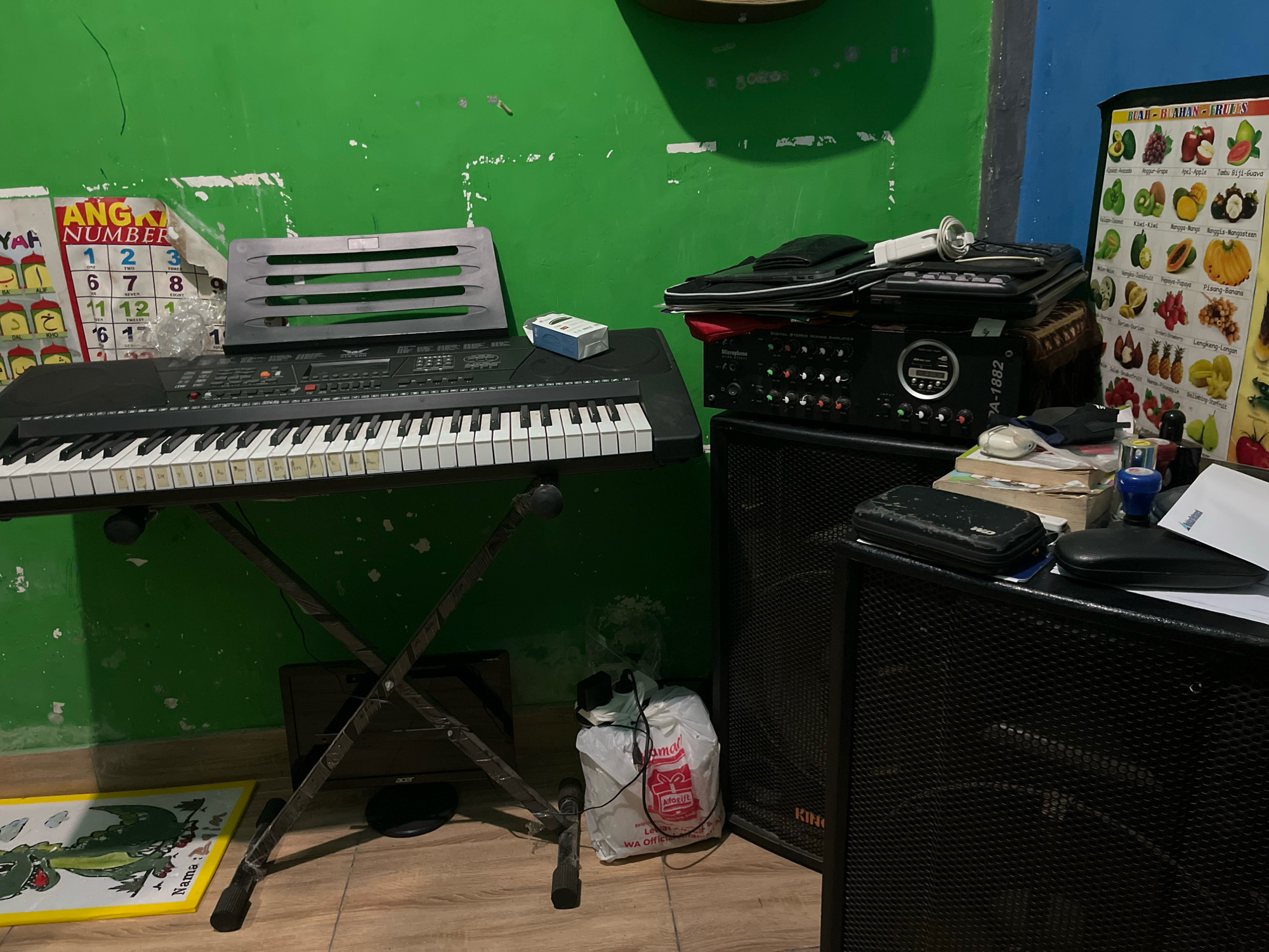 The image depicts an indoor scene, likely a room in Talang Kelapa, Indonesia, with a prominent electronic keyboard on a stand. The keyboard is black with white and black keys and is positioned in front of a green, distressed wall. To the left of the keyboard, a colorful educational chart with numbers and text is affixed to the wall. Above the keyboard, a black, plastic music stand rests. Further to the right, stacked on top of an audio amplifier, is a black bag containing what appears to be a microphone and other electronic accessories. A pack of cigarettes sits on the keyboard. The floor is wooden, and to the left, a yellow mat with a cartoon alligator illustration is visible. In the lower right portion of the image, a large black speaker cabinet is visible, with the "KING" logo faintly discernible. The overall impression is that of a personal music setup within a home environment.
