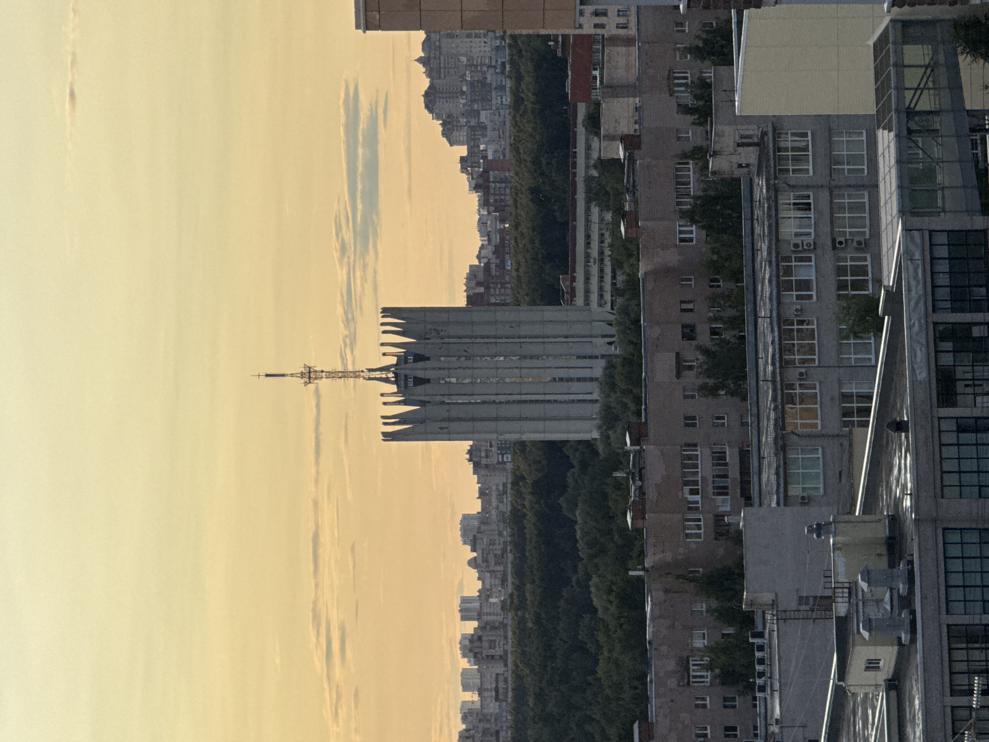 The image depicts a skyline view in an urban environment, likely during a sunset or sunrise given the warm hue of the sky and the soft lighting. In the foreground, several flat-roofed buildings are visible, some appearing to be industrial or commercial structures with large windows and ventilation systems on their roofs. These buildings are constructed with brick and concrete materials.
Dominating the mid-ground is a distinctive, tall, concrete building with a unique, pointed architectural style, resembling a crown or a series of fins at its apex. A radio tower or antenna is affixed to the very top of this structure. Behind this central building, a dense expanse of green trees forms a natural barrier, suggesting a park or forest within the city.
Further in the distance, a cluster of residential buildings of varying heights and architectural styles are visible, forming the city's silhouette against the horizon. The sky above is a pale yellow or orange, with scattered clouds adding texture to the scene. No people are visible in the image. The overall impression is a panoramic view of an urban landscape, characterized by a mix of modern and older architecture, industrial elements, and natural green spaces.