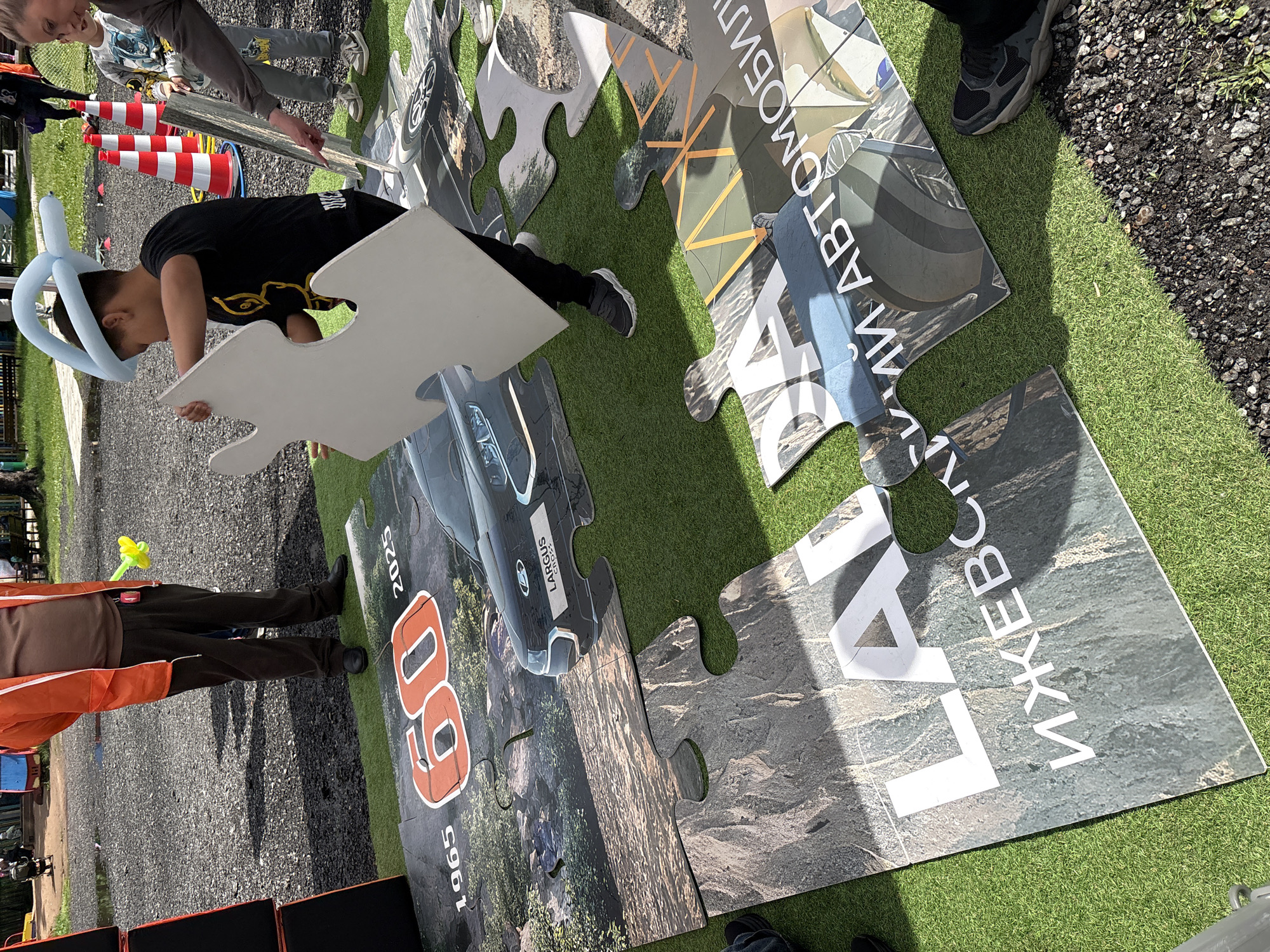 A boy with a light blue balloon on his head is holding a large, white puzzle piece in his hands, attempting to fit it into a large puzzle on the ground. The puzzle pieces display parts of cars, the numbers "60" with dates "1965" and "2025," and what appears to be the Russian Cyrillic text "Lada Izhevsk Avtomobili." The puzzle is laid out on a patch of green artificial turf. Two other people, partially visible in the frame, appear to be involved in the activity. Traffic cones are visible. The location is Izhevsk, Russia.