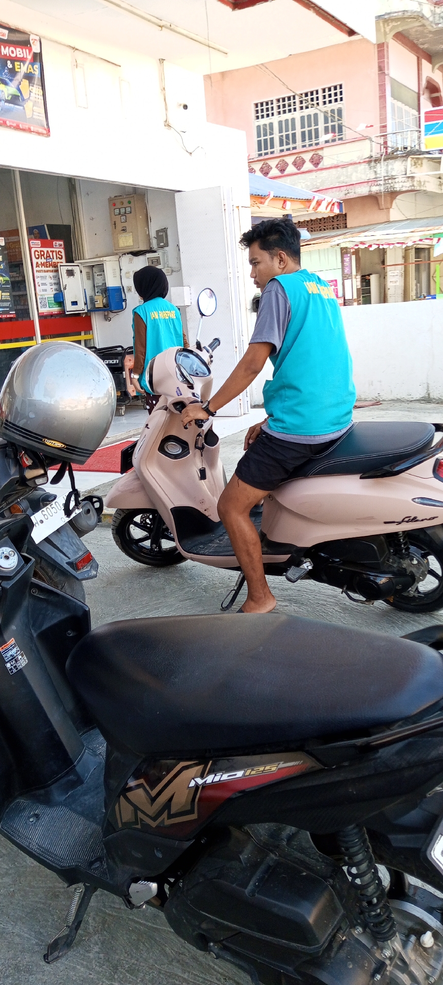 In the city of Assorajang, Indonesia, two individuals are depicted at what appears to be a minimart or a roadside convenience store. The scene is captured from a low angle, focusing on a light beige scooter with a dark seat. A young man, wearing a turquoise vest over a gray t-shirt and black shorts, is seated on the scooter, looking to his left. His bare feet are visible. Behind him, another person, dressed in a black headscarf and a turquoise vest identical to the man's, stands near a counter or a service area. In the foreground, another scooter, predominantly black, is partially visible, with its seat in focus. The background reveals a building with a pink facade, characterized by windows with white grid-like structures and a red and white striped awning. Signs with Indonesian text are present on the building, suggesting commercial activity. The environment appears to be outdoors, with concrete flooring.