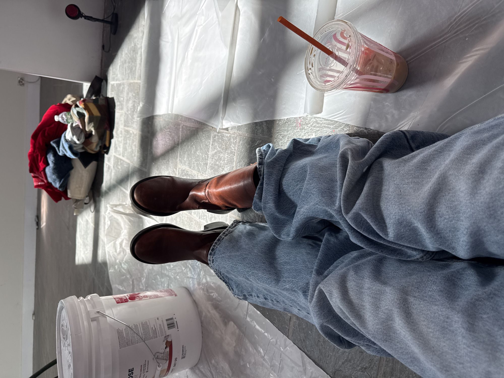 The image shows a person sitting on a plastic drop cloth wearing blue jeans and brown boots. A white bucket is to the left, and a clear plastic cup with a straw is to the right. In the background, a pile of clothes and a desk lamp are visible. The location appears to be indoors, possibly a room being prepared for painting or renovation. The city of Town of Red Hook, United States of America is only a best guess. No other specific details are available to pinpoint an exact location within the city.