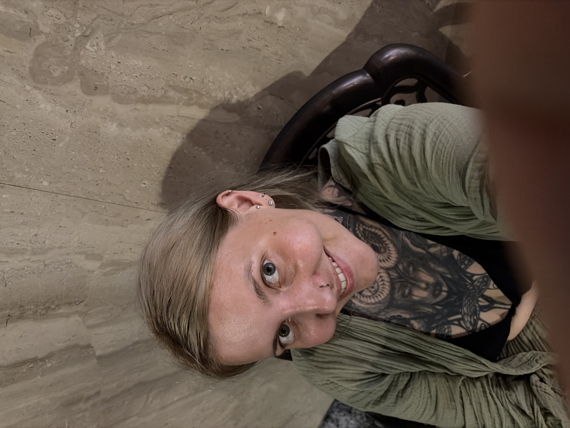 A close-up, high-angle portrait captures a young adult female smiling brightly and looking directly upwards at the camera. Her head is tilted, giving the image an inverted perspective. She has light-colored hair, likely blonde or light brown, framing her face. Her left ear is adorned with multiple silver piercings, including a helix, conch, and lobe piercings. A silver septum ring is visible on her nose. A prominent, intricate black tattoo covers her left shoulder and upper chest, depicting a detailed design that appears to include a face or mythological figure, possibly with snakes or flowing elements. She is wearing a light green, textured, crinkled fabric top or jacket over a dark, possibly black, inner garment.
The background consists of a light beige or grey textured floor, resembling marble or tile, with natural stone patterns and specks. Behind her head and shoulder, a dark, curved piece of furniture, likely a chair, is partially visible, casting shadows on the floor. The lighting appears ambient and somewhat subdued. A blurred, out-of-focus element, possibly a hand or finger, is visible on the right edge of the frame. The woman's expression is one of clear joy and engagement. The image is set in Đà Nẵng, Vietnam. There is no visible text.