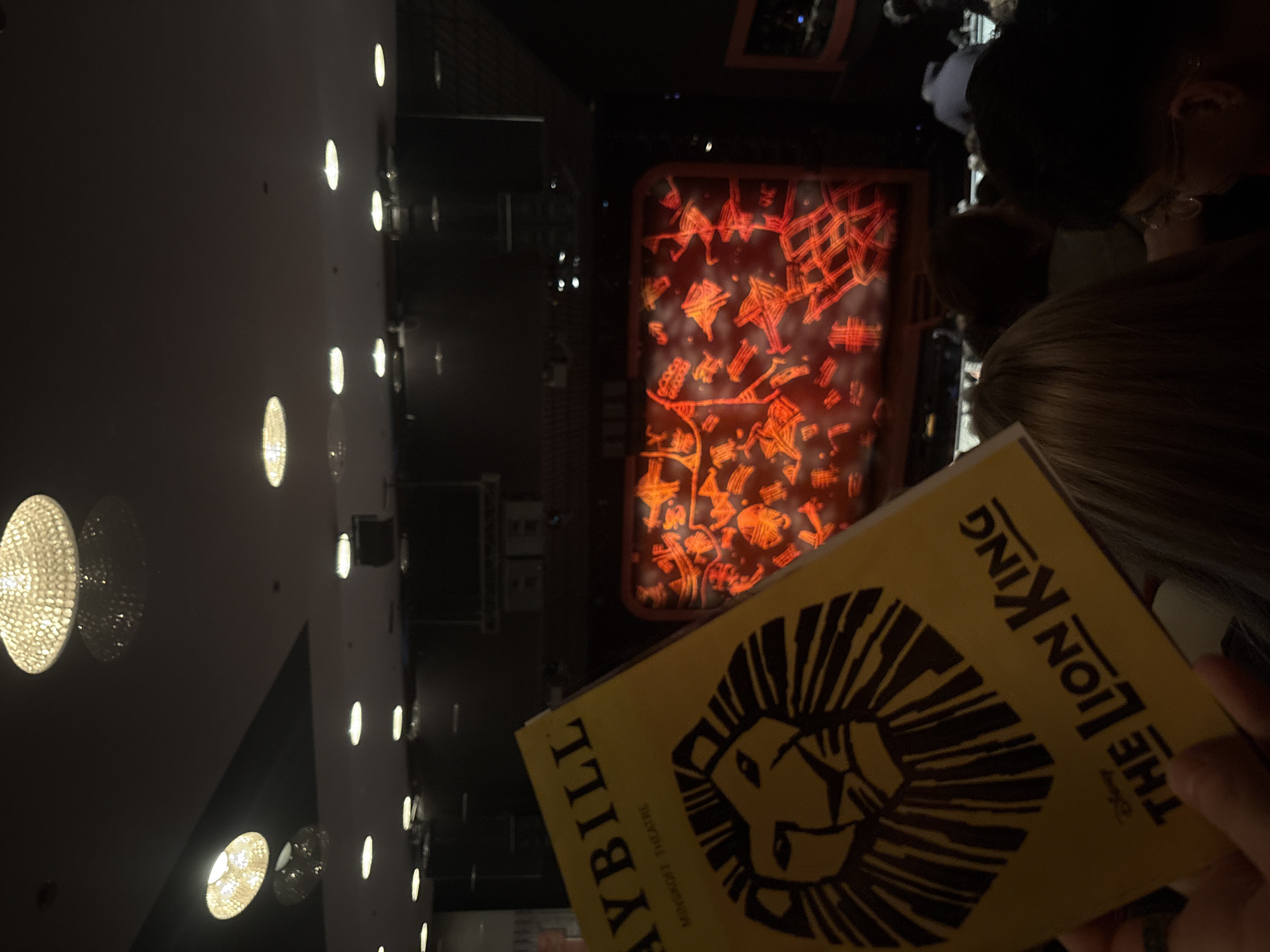 The image shows a person holding a playbill for "The Lion King" at the Minskoff Theatre in New York City. The playbill features a yellow cover with a black silhouette of a lion's head. In the background, a brightly lit stage is visible, with a red and orange design. Other people are sitting nearby. The environment appears to be dimly lit, with rows of lights visible on the ceiling.