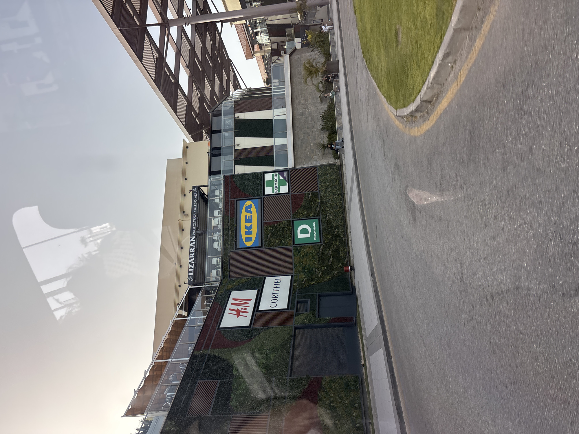 A side view of a modern shopping center is captured in this image, located in Urbanización La Cenia II, Spain. The building features a prominent green wall adorned with various store signs, including H&M, Cortefiel, IKEA, and Deichmann. Further up, a sign reads "LIZARRAN PINCHOS, TAPAS Y MUCHO MÁS." A portion of the building has a distinctive cantilevered roof structure. In front of the building, a paved walkway runs alongside a grassy area. Several individuals are seated on a low wall in front of the building, while others are standing near the entrance to another section of the shopping center, which has a sign that reads "Tapas & Beer." The scene is framed by a road with a yellow line marking the edge, curving towards the right side of the image.