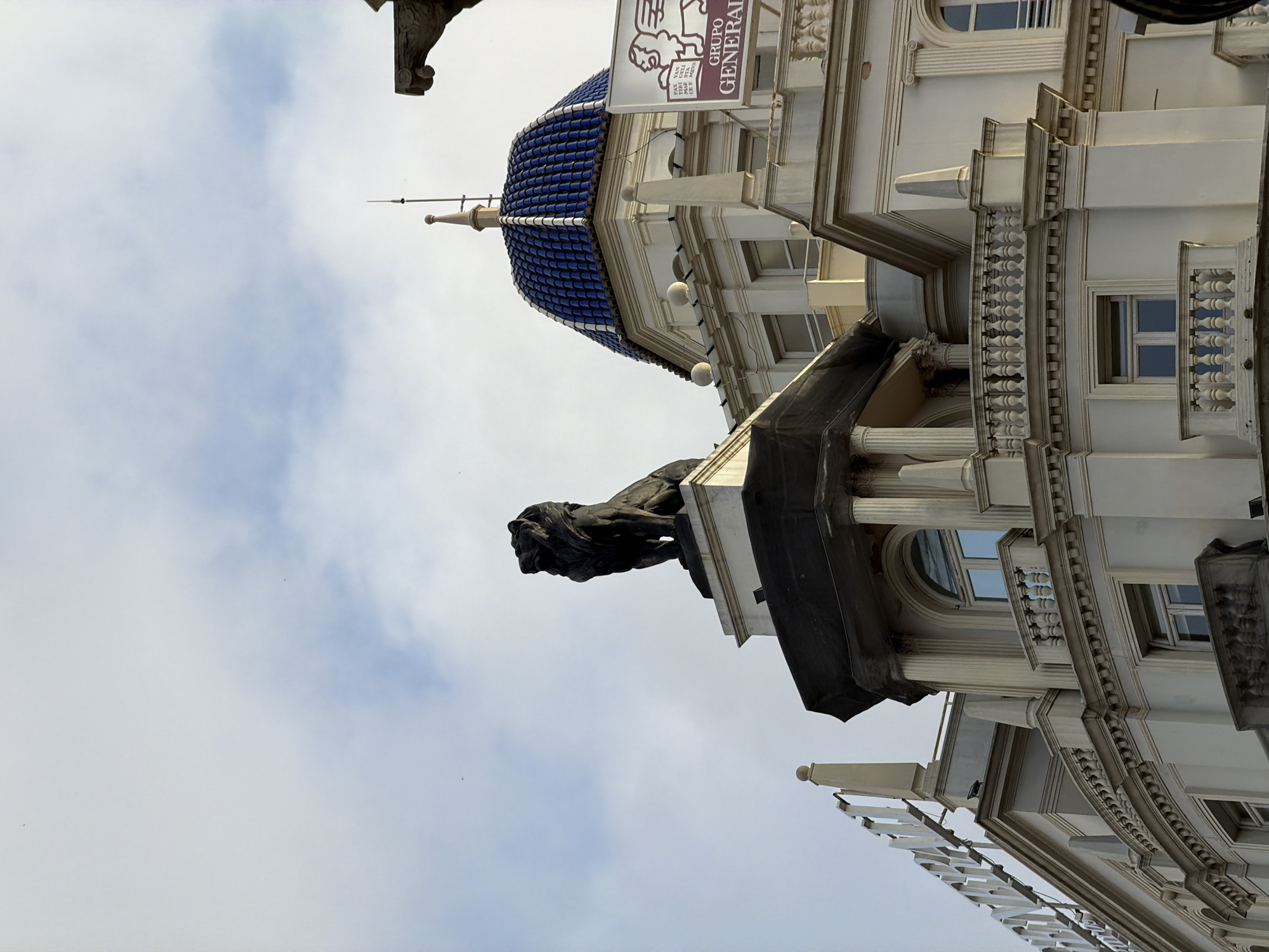 The image shows a close-up, low-angle view of the upper facade of an ornate, light-colored building against a partially cloudy sky. A dark, statue of a lion is prominently displayed on a pedestal atop a section of the building, which is partially covered by a black tarp. To the right, a blue tiled dome with a finial rises from the building. A sign with the text "GRUPO GENERALI" and a winged lion logo is attached to the building. Architectural details such as columns, balconies with balustrades, and arched windows are visible. In the upper left corner of the frame, a partial view of a large sign with white lettering can be seen. The overall impression is of grand, possibly historical, architecture in an urban setting.